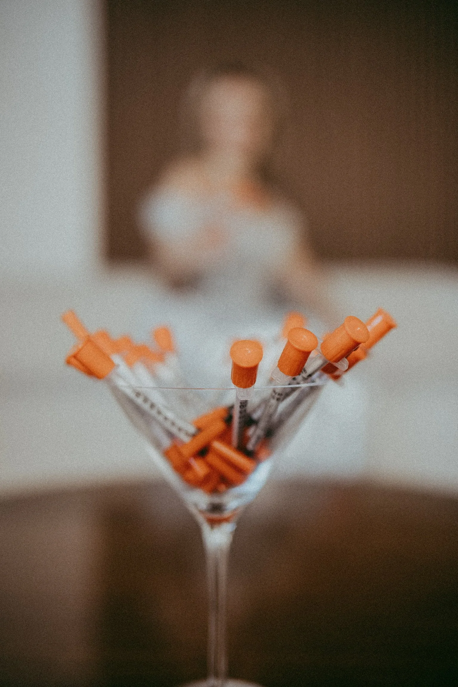 A vertical shot of a clear drink with orange liquid-filled syringes arranged around the rim of the glass. The background shows an out-of-focus person.