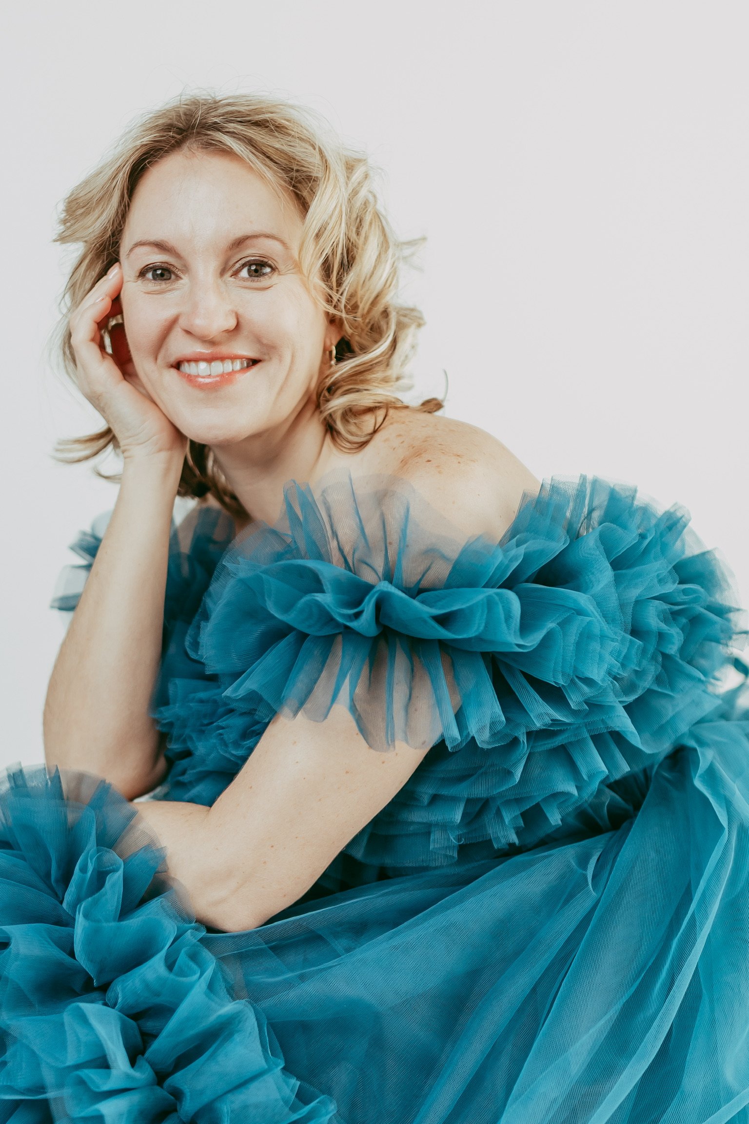 A woman with blonde, wavy hair smiling, resting her head on her left hand, wearing a blue, ruffled dress against a white background.