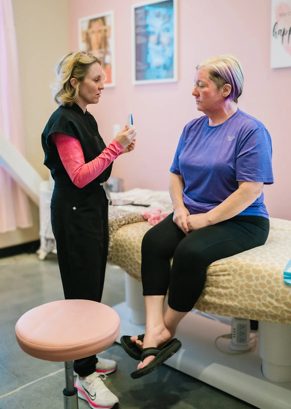 A healthcare professional points to a test strip or small device while talking to a woman sitting on an examination table in a medical room. The woman is listening attentively, sitting with her legs crossed and hands resting on her lap. The room has pink walls and framed pictures.