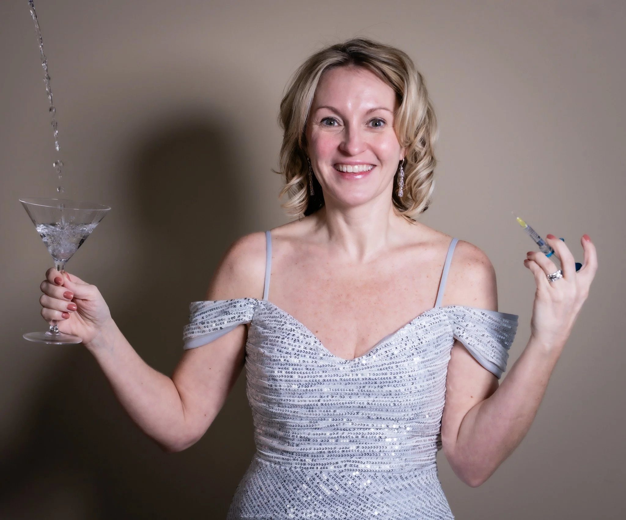 A smiling woman in a shiny silver dress holding a martini glass in her right hand and a syringe in her left hand. The background is plain, and she has curly blonde hair, earrings, and a ring on her left hand.
