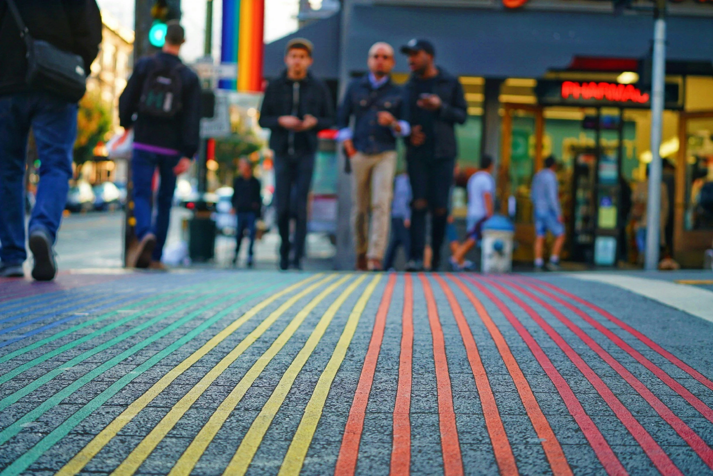 Colorful rainbow-striped crosswalk on a busy city street with pedestrians walking and talking in the background.