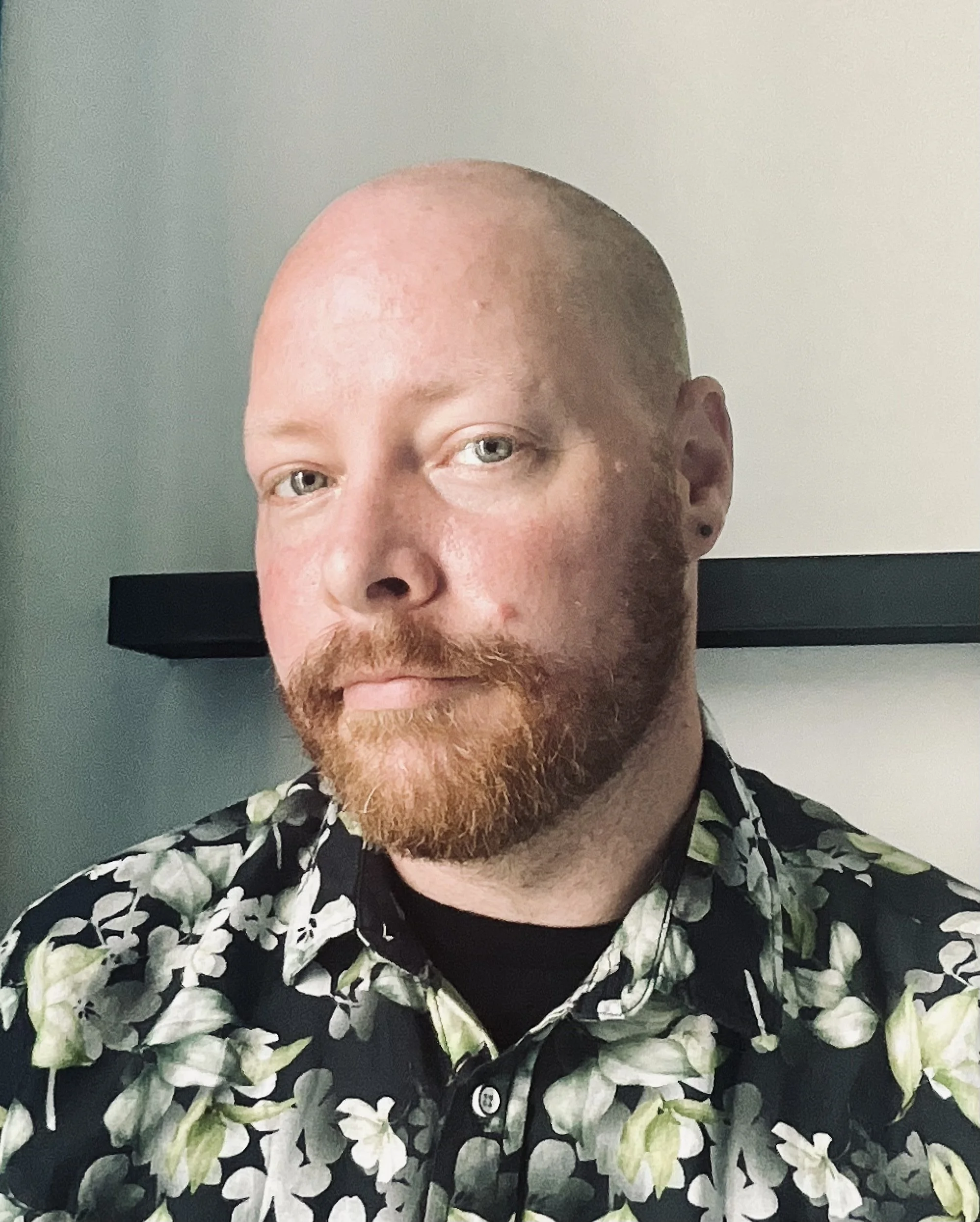 A person with a shaved head and a reddish beard wears a shirt with a floral pattern.