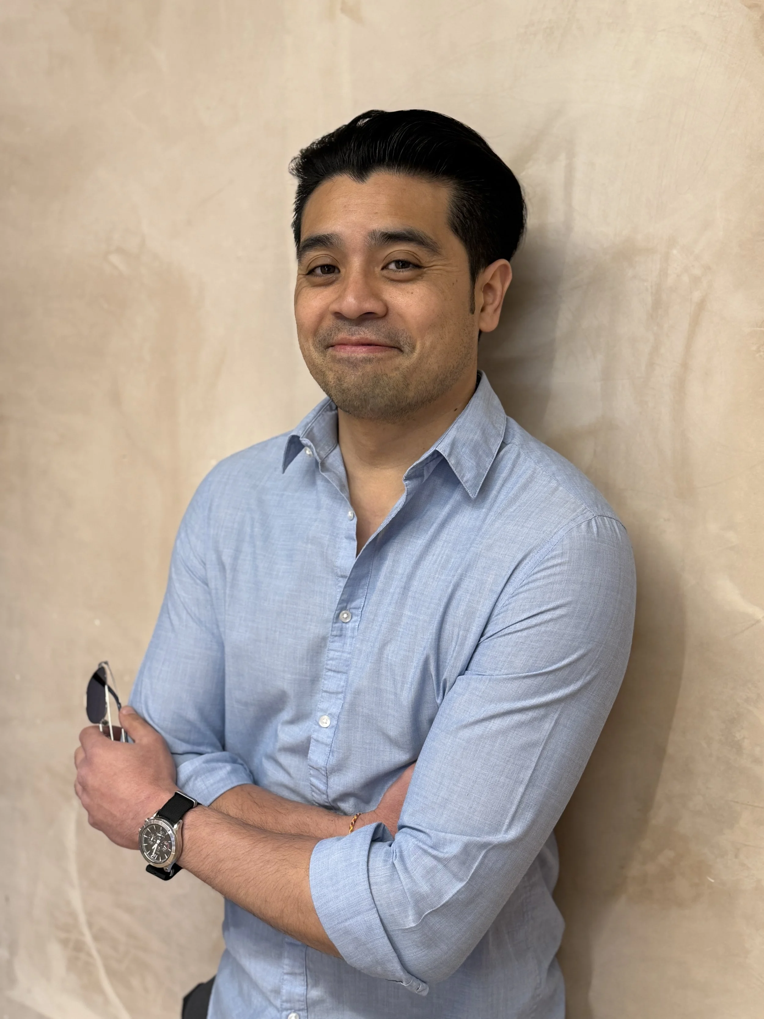 A man with dark hair, wearing a light blue button-up shirt, standing against a beige wall, smiling at the camera, holding glasses in his right hand and wearing a watch on his left wrist.