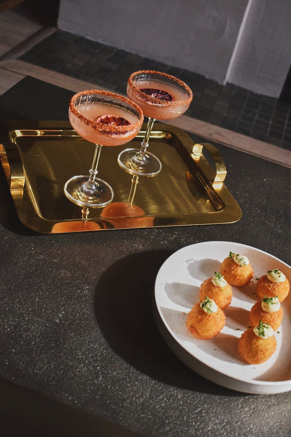 Two cocktails with salted rims and cherry garnishes on a gold tray, and a plate of six fried appetizers topped with sauce, all on a black table.