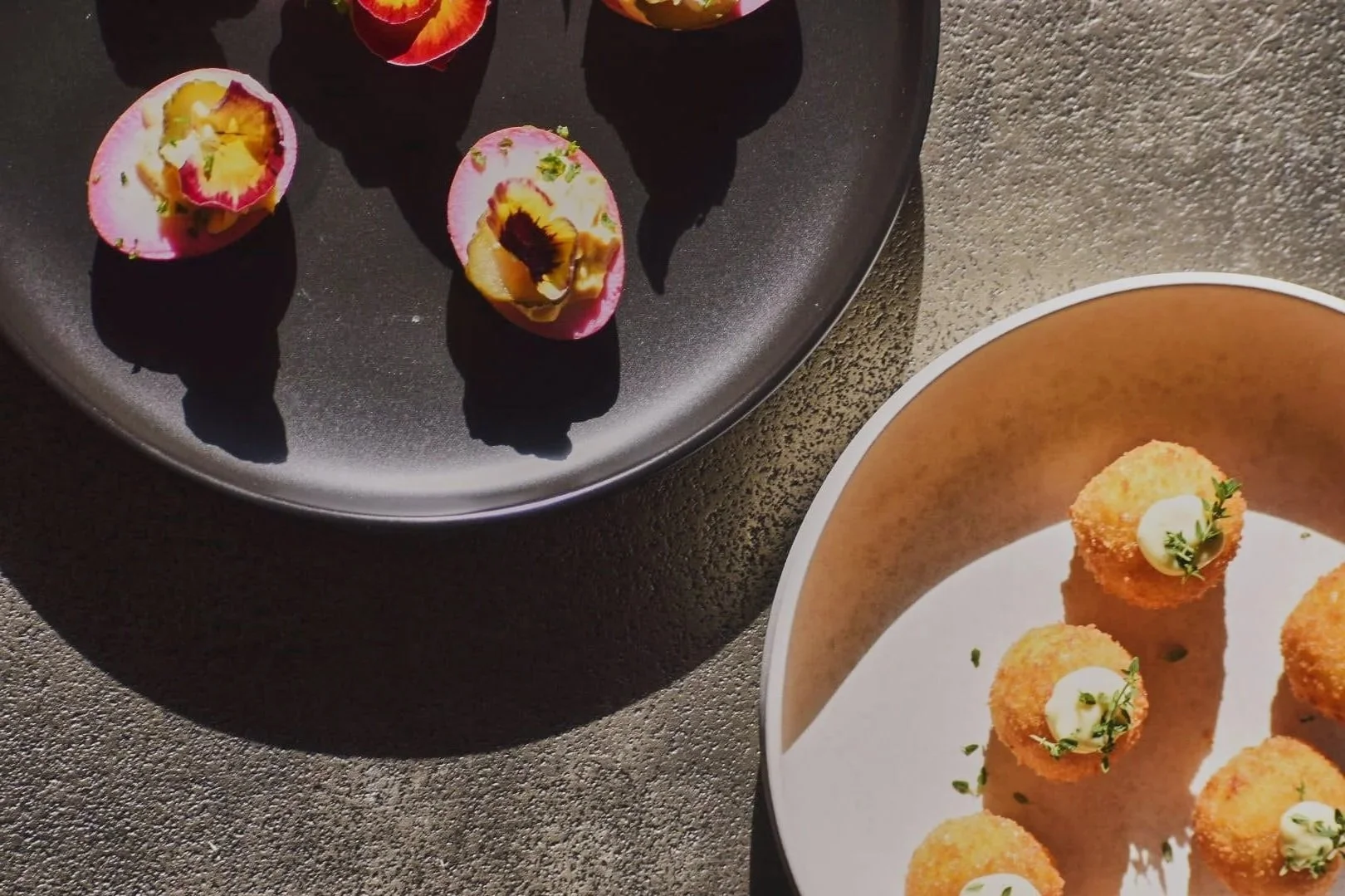 Close-up of two plates with appetizers: one black plate with halved pink and yellow passion fruit, and one beige plate with breaded, fried bite-sized foods topped with small dollops of sauce and sprigs of herbs.