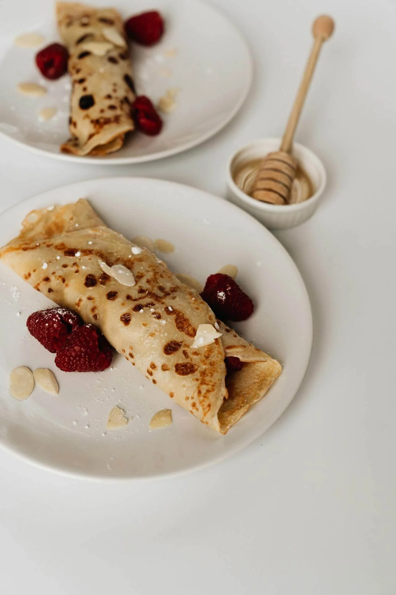 Two white plates with crepes topped with raspberries and almond slices, honey jar with a honey dipper, on a white surface.
