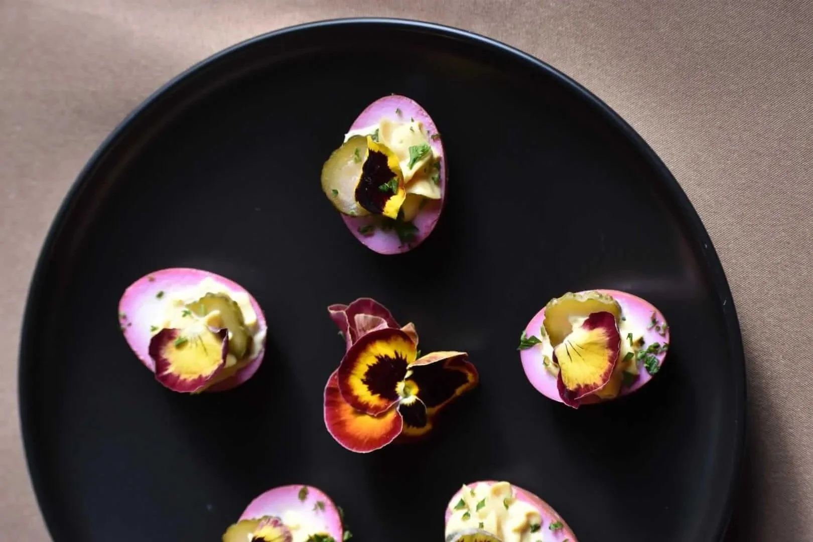 Elegant black plate with five pink-colored appetizers topped with herbs, edible flowers, and small garnishes, arranged around the center with scattered flower petals.