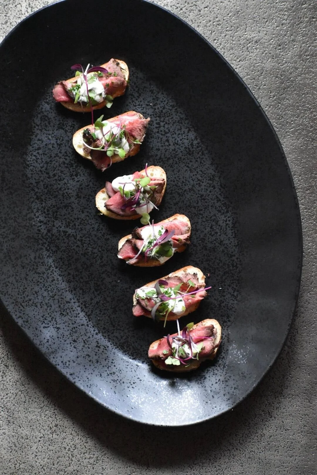 Seven pieces of sliced beef tenderloin on toasted baguette slices garnished with microgreens and a drizzle of white sauce on a black oval plate.