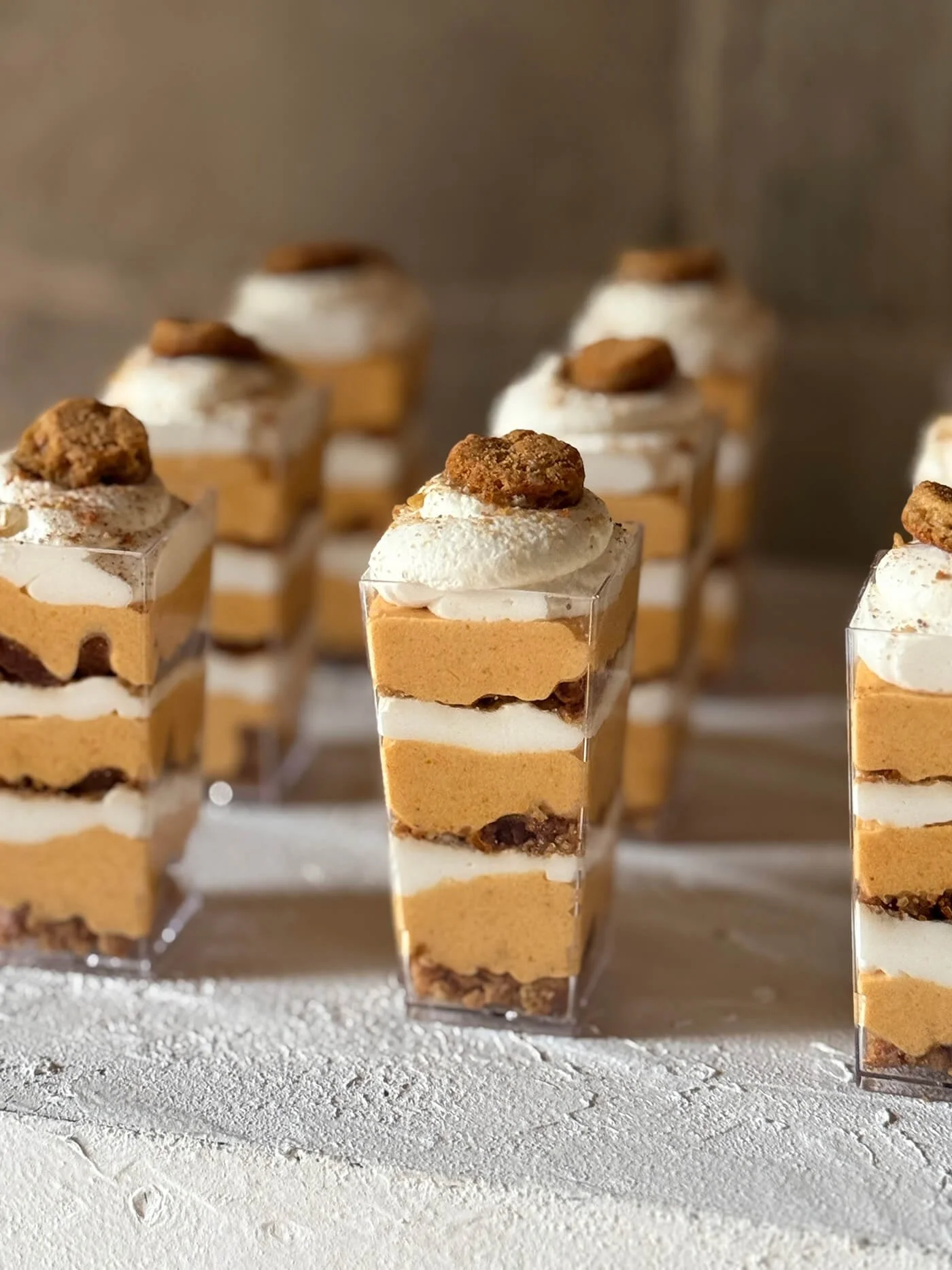 Multiple layered desserts in clear cups, topped with whipped cream and cookie pieces, on a textured white surface.