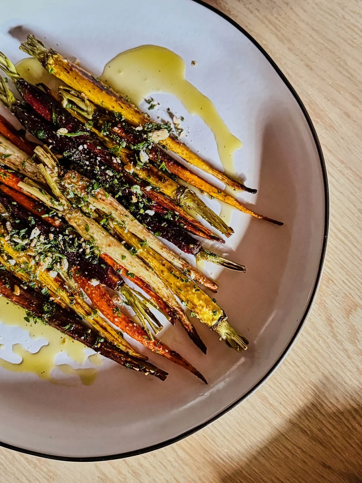 A plate of roasted colorful carrots drizzled with olive oil and garnished with chopped herbs, on a white marble or ceramic dish with a black rim.