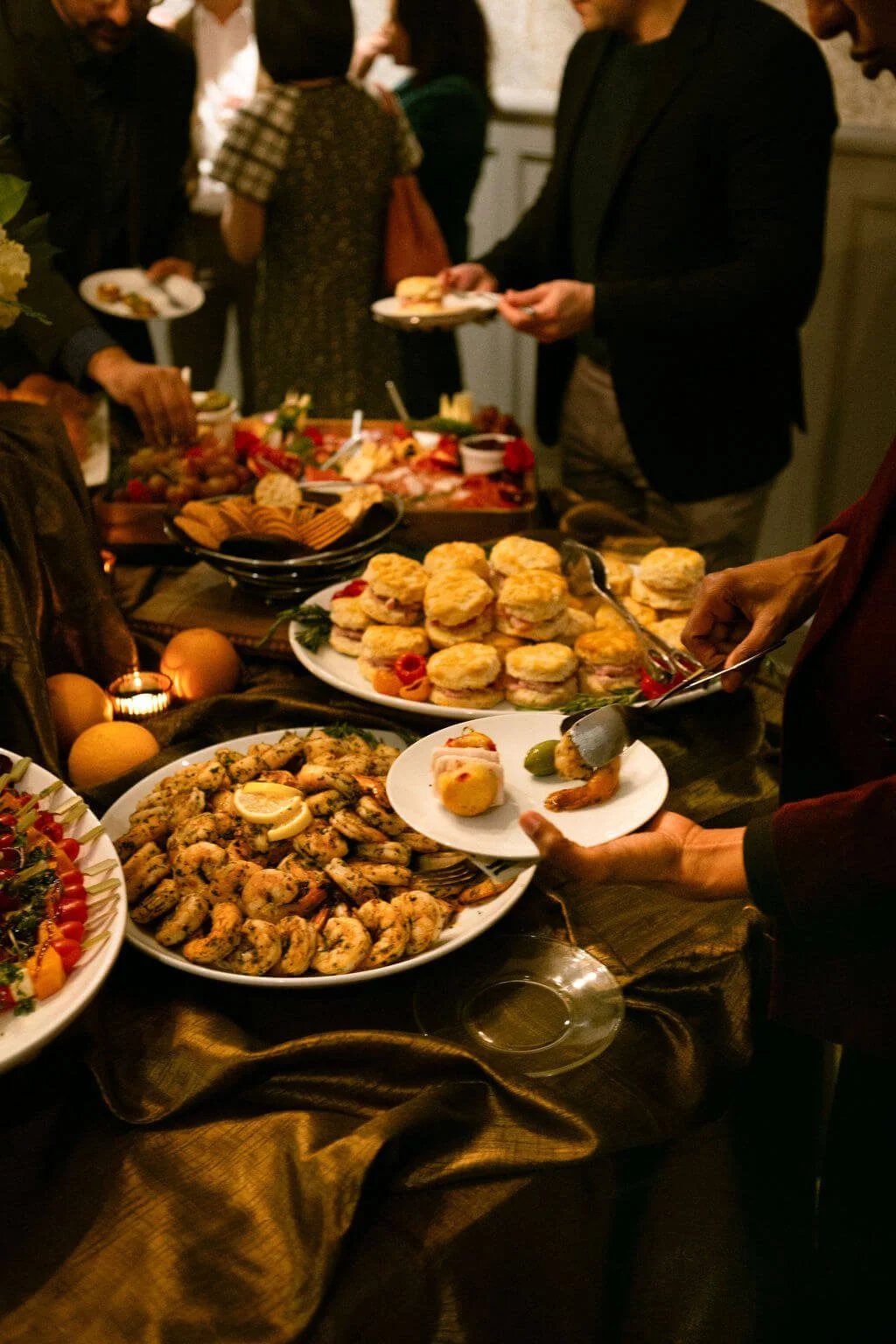 People serving themselves food at a buffet table with various dishes including sandwiches, shrimp, salads, and desserts.