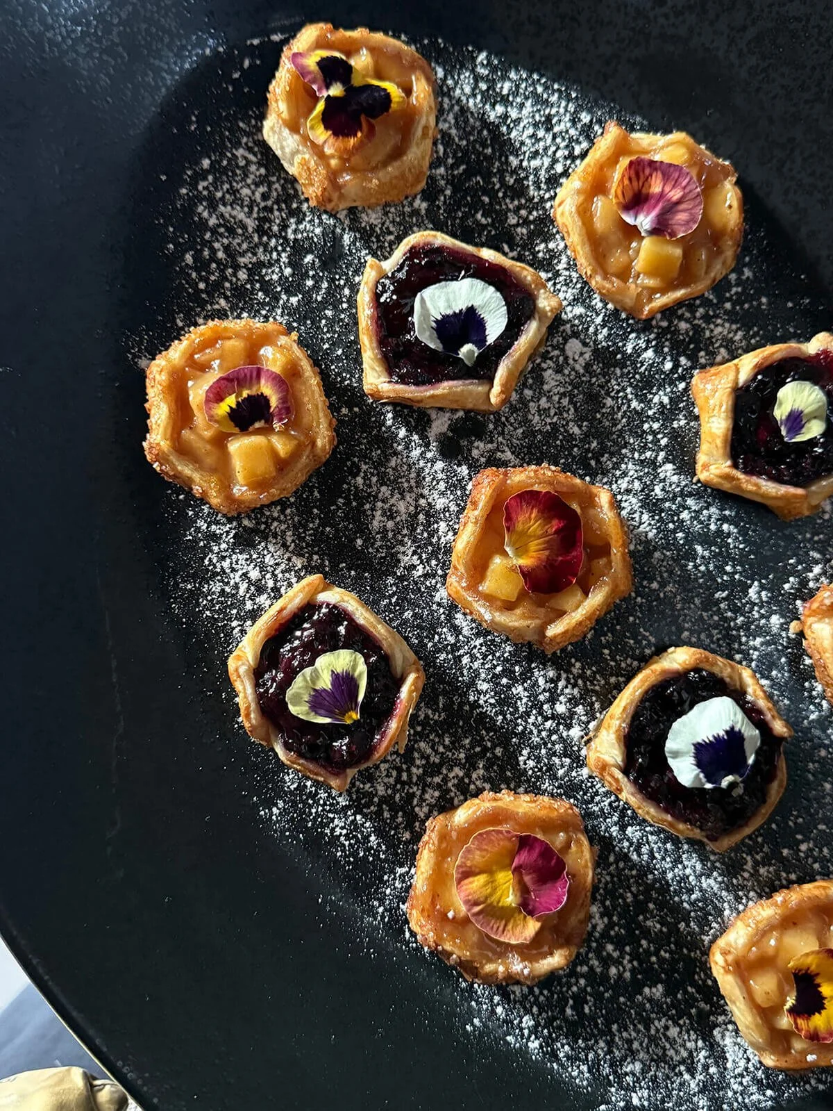 Assorted mini pastries topped with edible flower petals, arranged on a black plate dusted with powdered sugar.