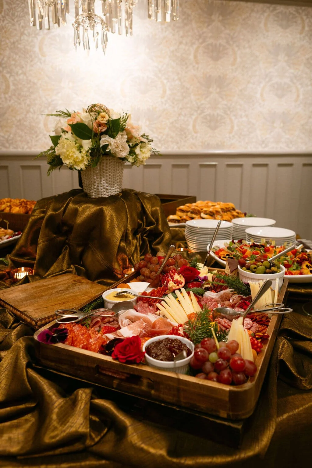 Buffet table with cheese, grapes, cold cuts, and salads, decorated with a flower arrangement atop a gold-colored cloth in an elegant dining room.