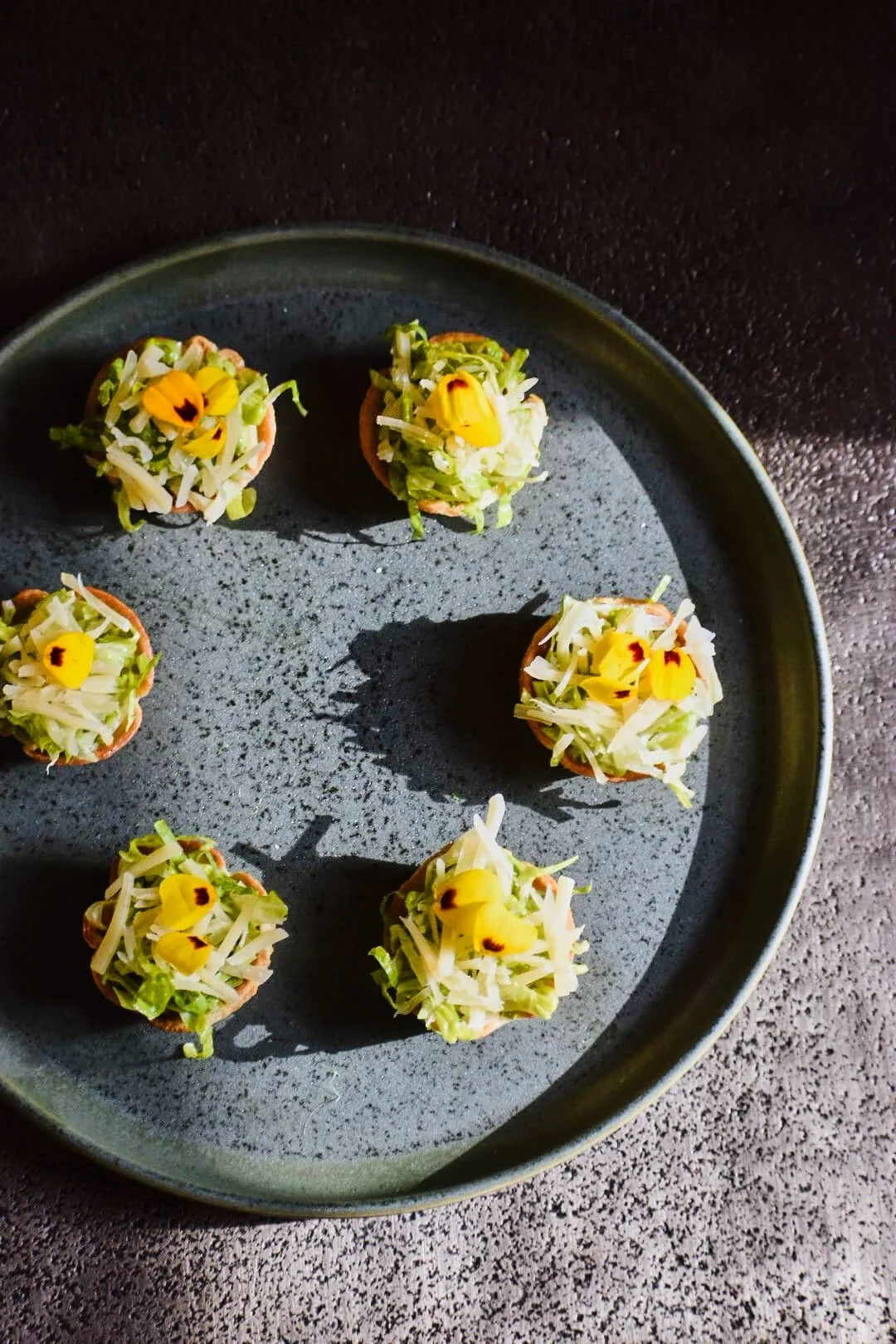 Small appetizers topped with shredded cheese, lettuce, and edible flowers on a dark gray plate.