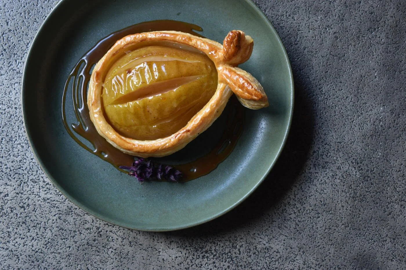 A tart with a whole lemon inside, topped with caramel glaze, served on a green ceramic plate with a drizzle of caramel and a small purple edible flower.