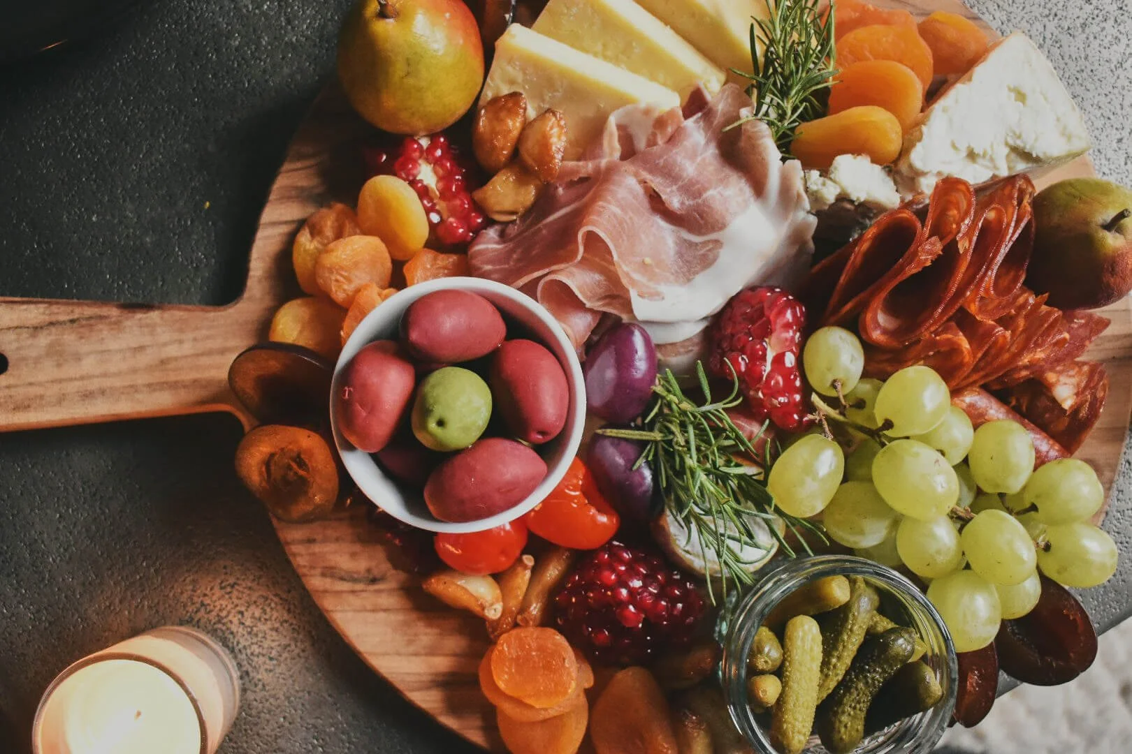 A cheese and charcuterie board with grapes, dried apricots, a bowl of olives, cured meats, cheese slices, and garnished with rosemary, on a wooden serving platter.