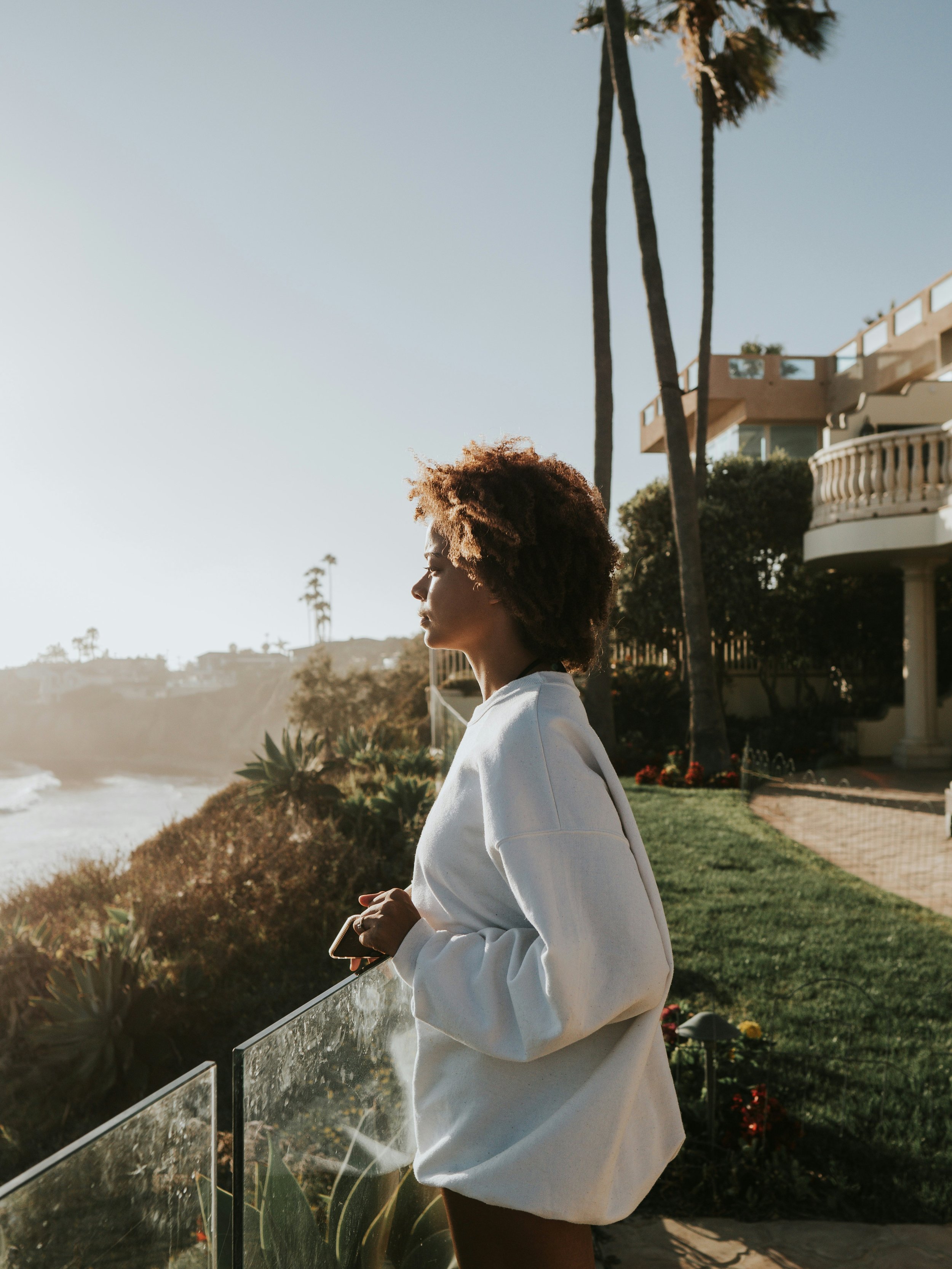 A woman with curly hair walking outdoors near a coastline with palm trees, buildings, and an ocean sunset in the background.