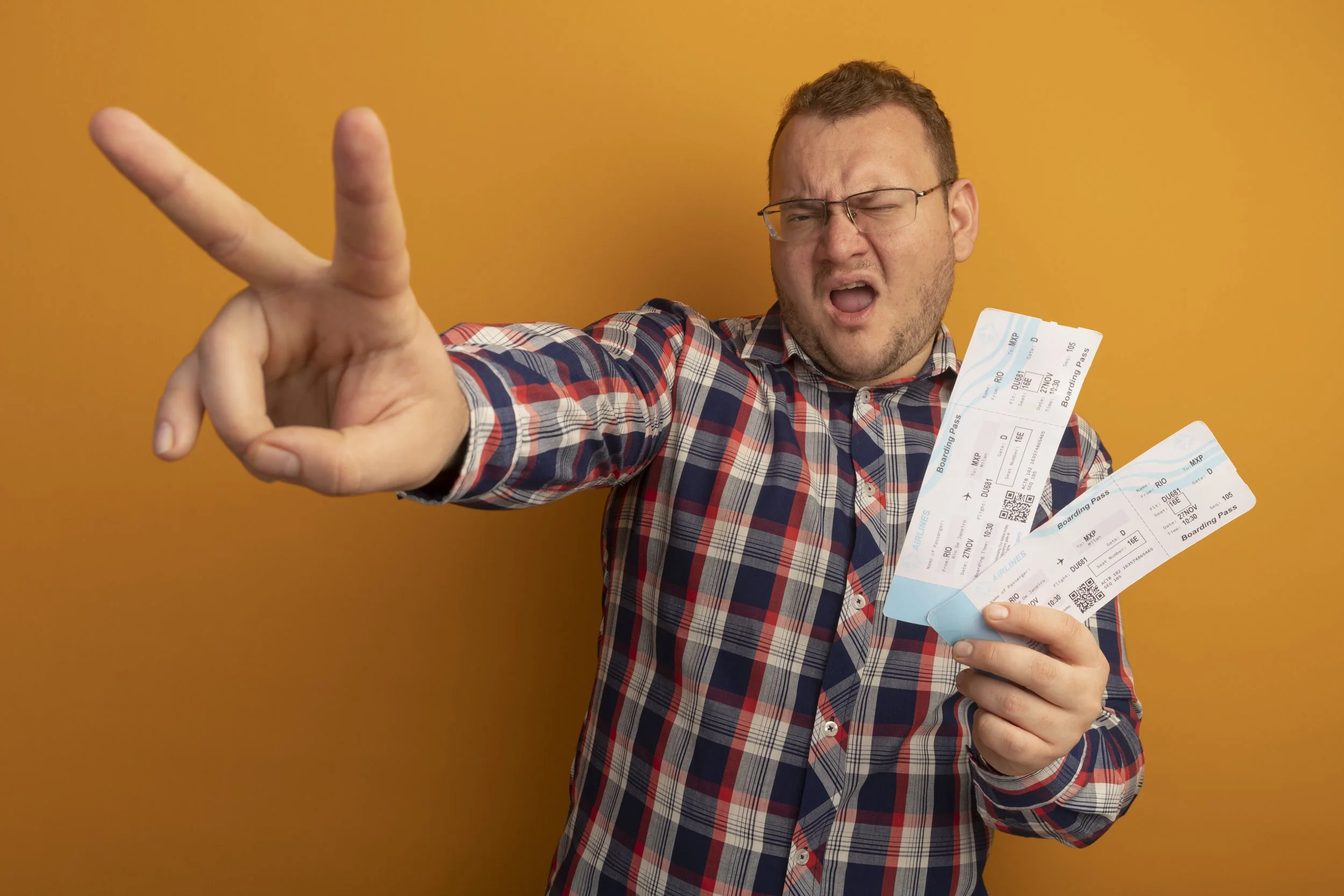 emotional-man-glasses-checked-shirt-holding-air-tickets-looking-aside-showing-v-sign-standing-orange-wall.jpg