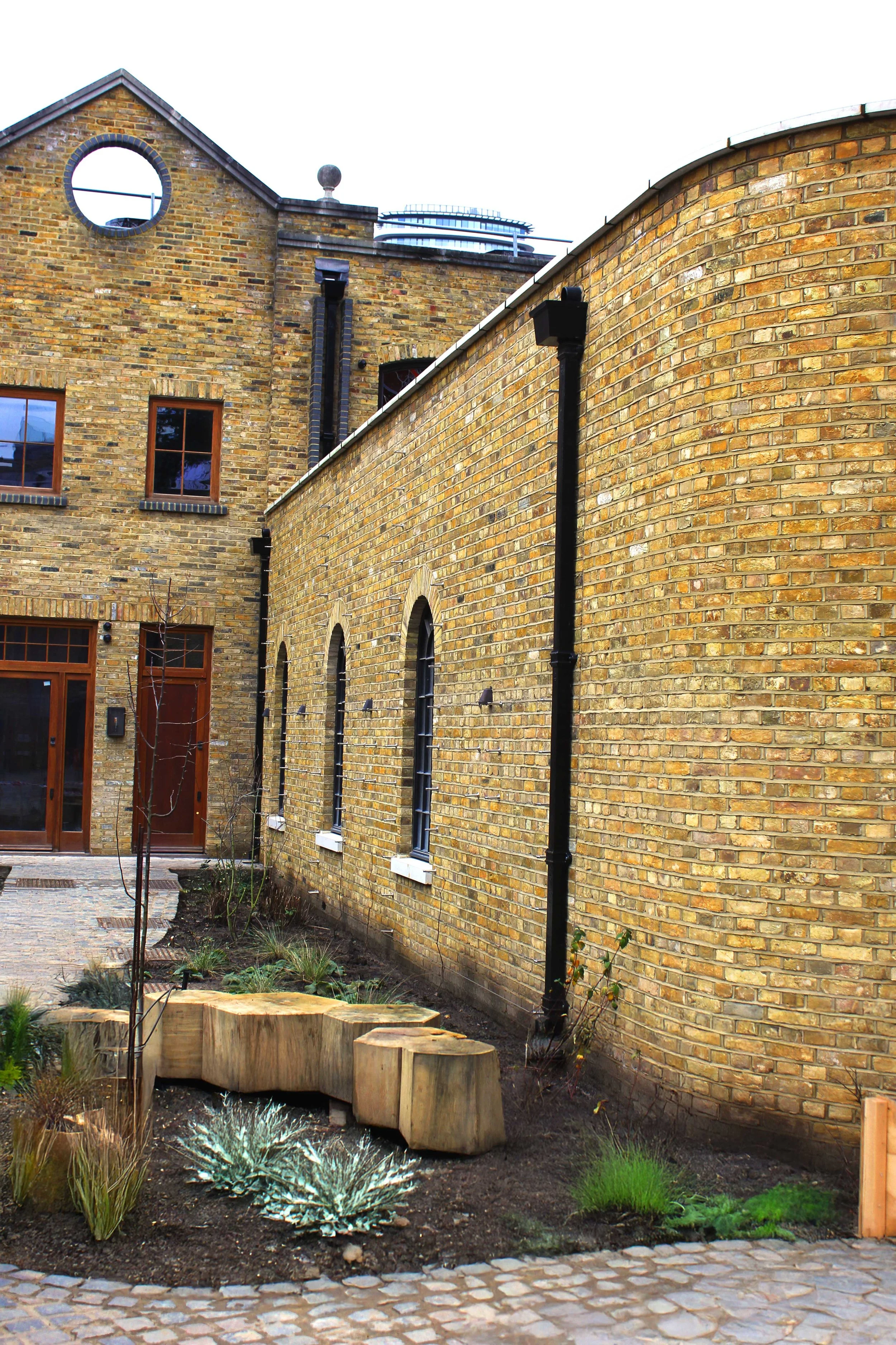 View of a yellow brick residential building with arched windows and black rain gutters, adjacent to a small garden area with young plants and wooden logs.