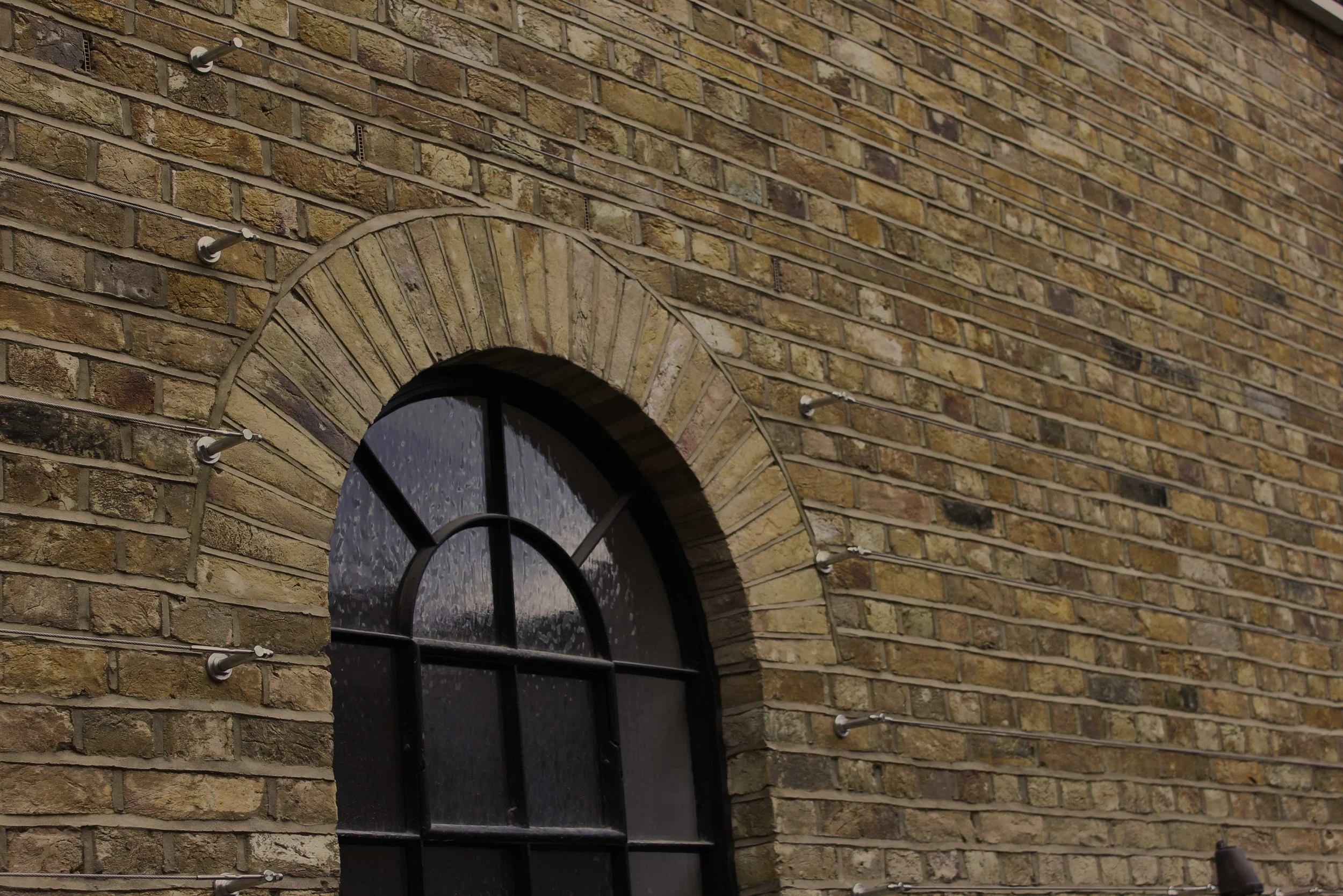 Close-up of a black arched window with raindrops on the glass, set into a yellow and brown brick building. There are several metal hooks and wires attached to the brick wall around the window, and a small outdoor light fixture at the bottom right corner.
