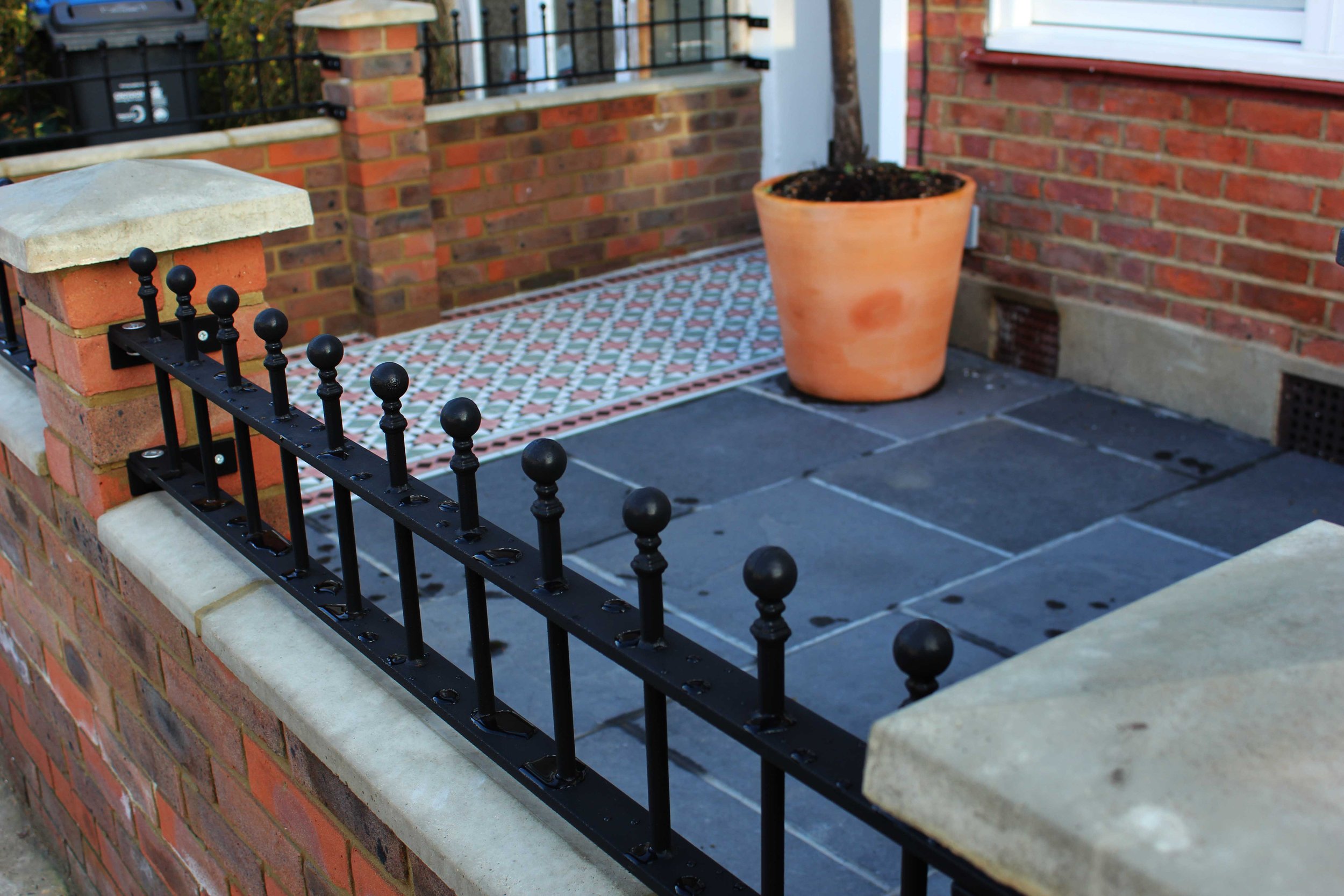 A small outdoor patio area with black iron railing, blue paving stones, a large terra cotta plant pot with soil and a tree, and a brick wall with a window.