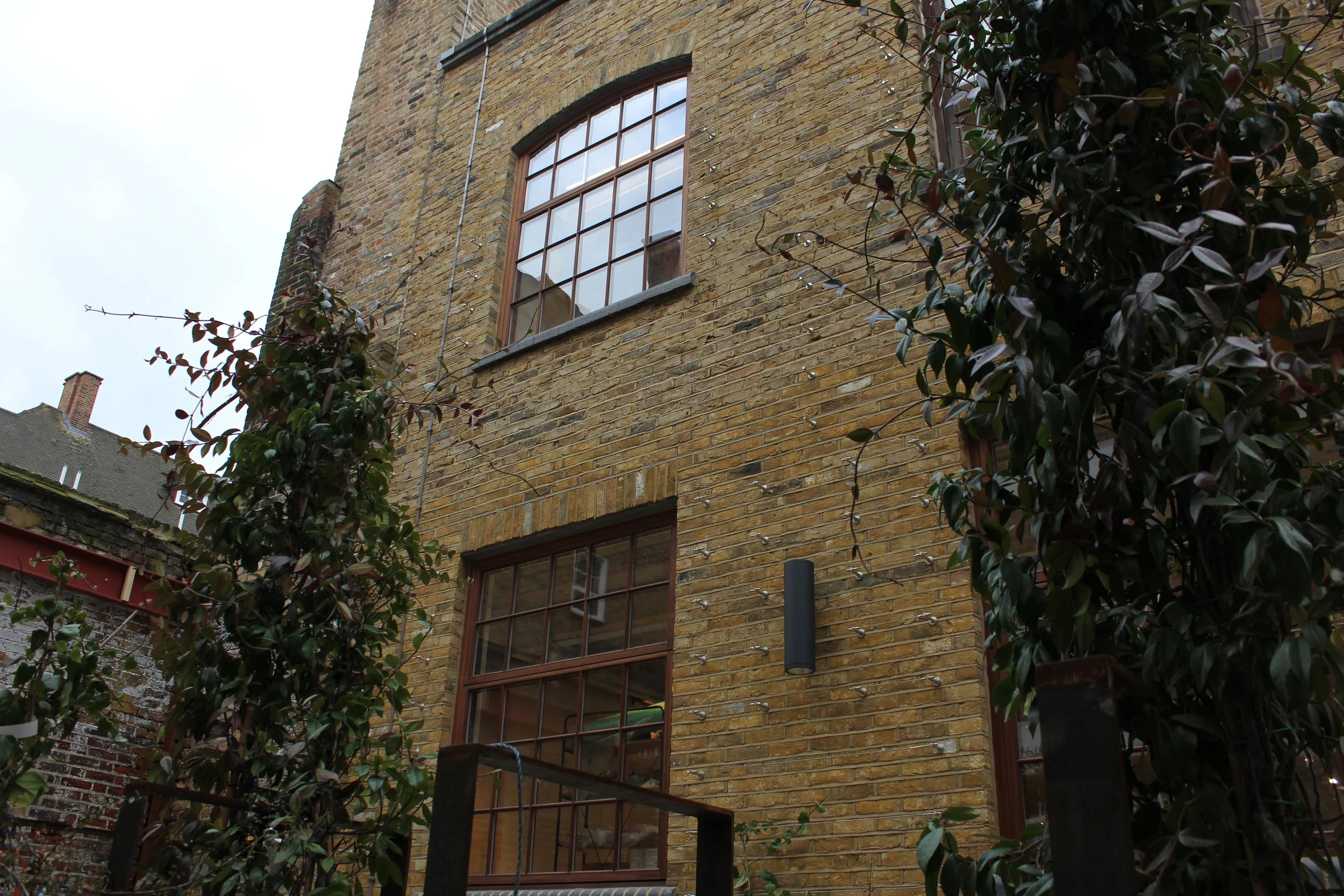 Close-up view of a brick building with large brown-framed windows and greenery on the side, under an overcast sky.