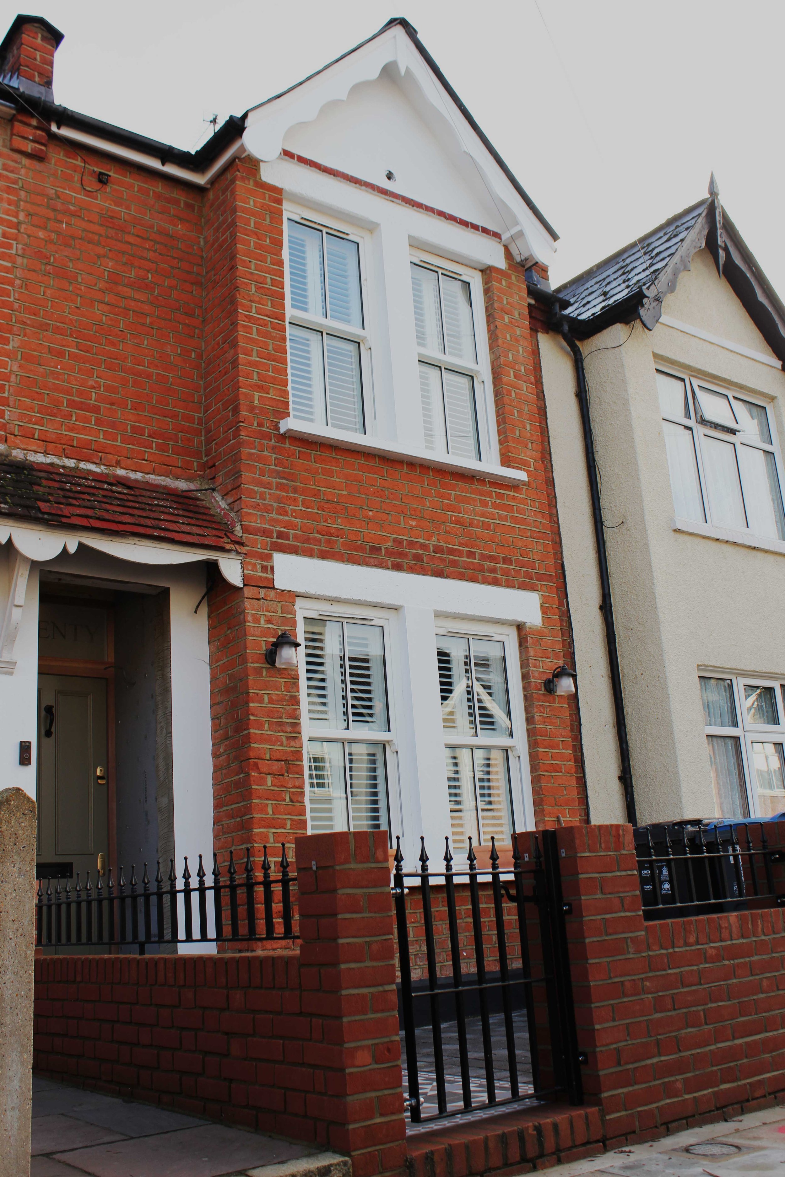 A red brick house with white trim and large windows with white shutters, a small front yard with a brick and black metal fence, and a neighboring cream-colored house.