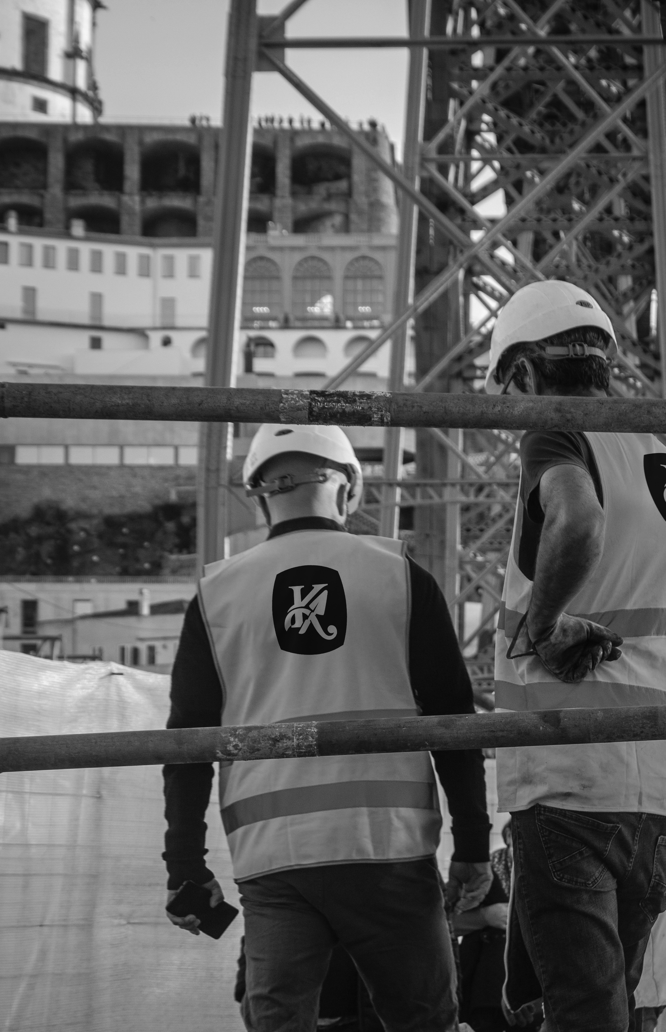 Two construction workers wearing hard hats and safety vests on a construction site, with an unfinished building and metal scaffolding in the background.