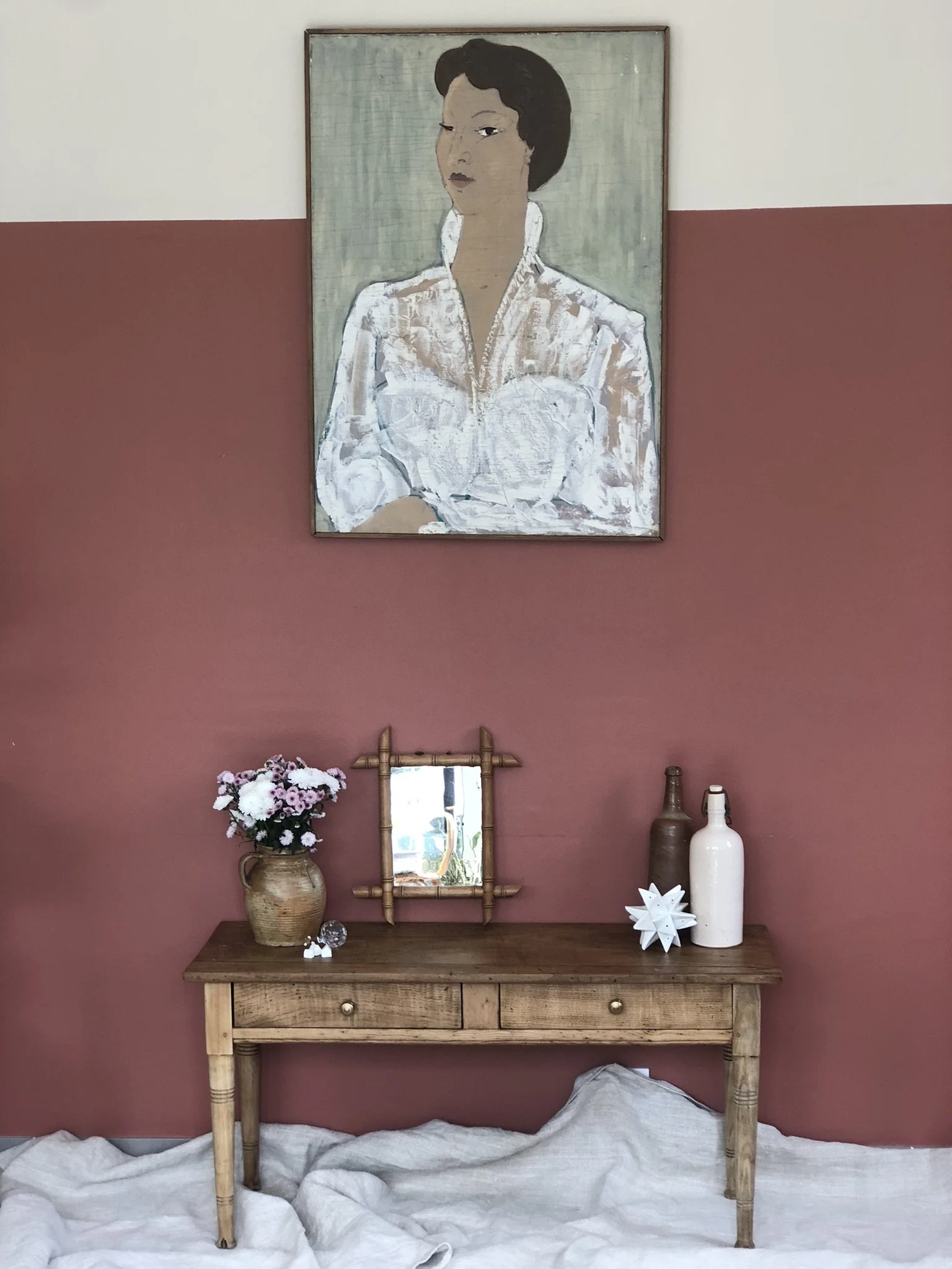 Une console en bois avec des vases et des décorations, un miroir en bambou et un tableau d'une femme avec une chemise blanche