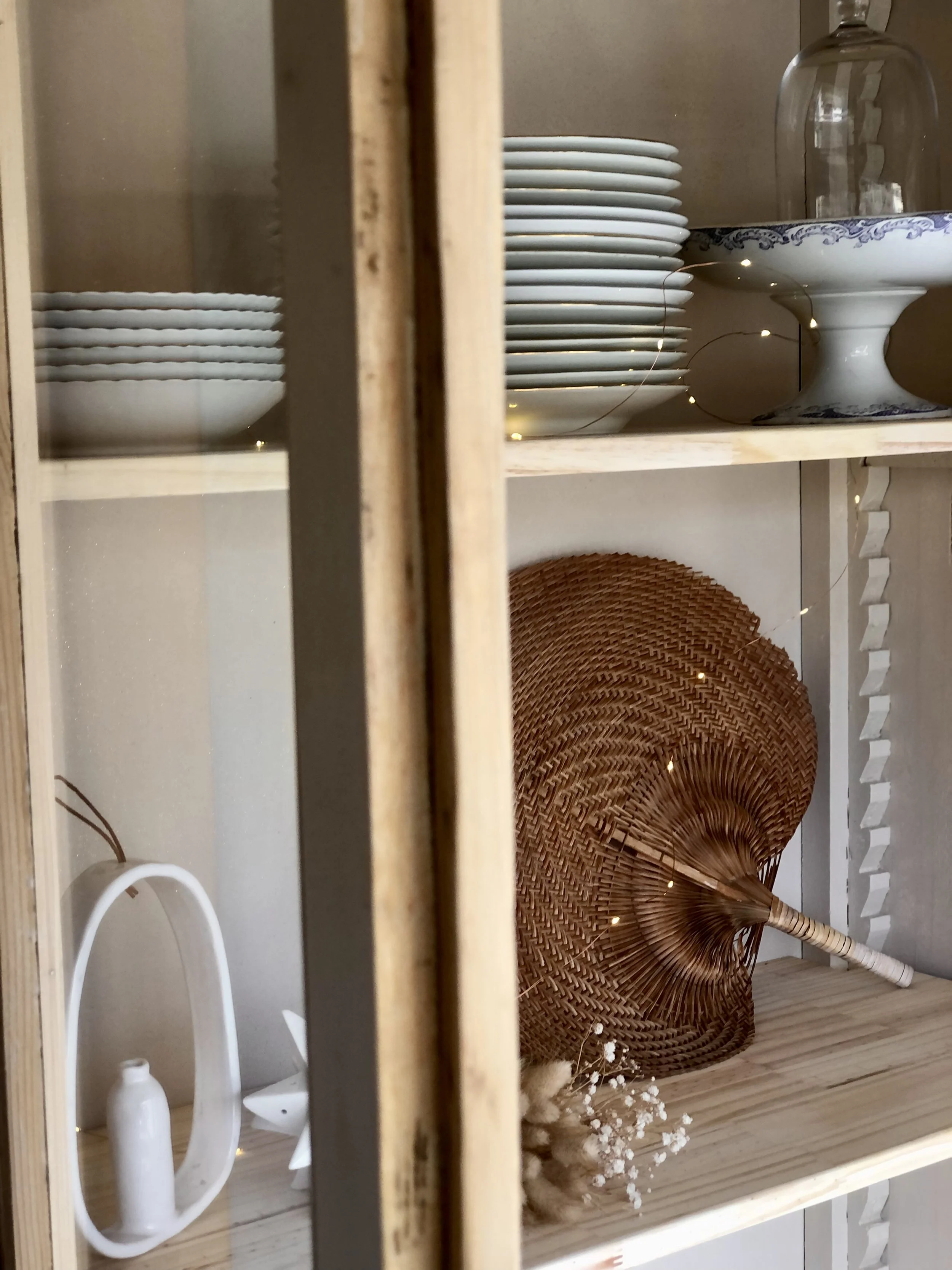 Une étagère en bois clair contenant des assiettes blanches empilées, une coupe avec un motif bleu, quelques décorations en fibres naturelles et une bouteille en verre.