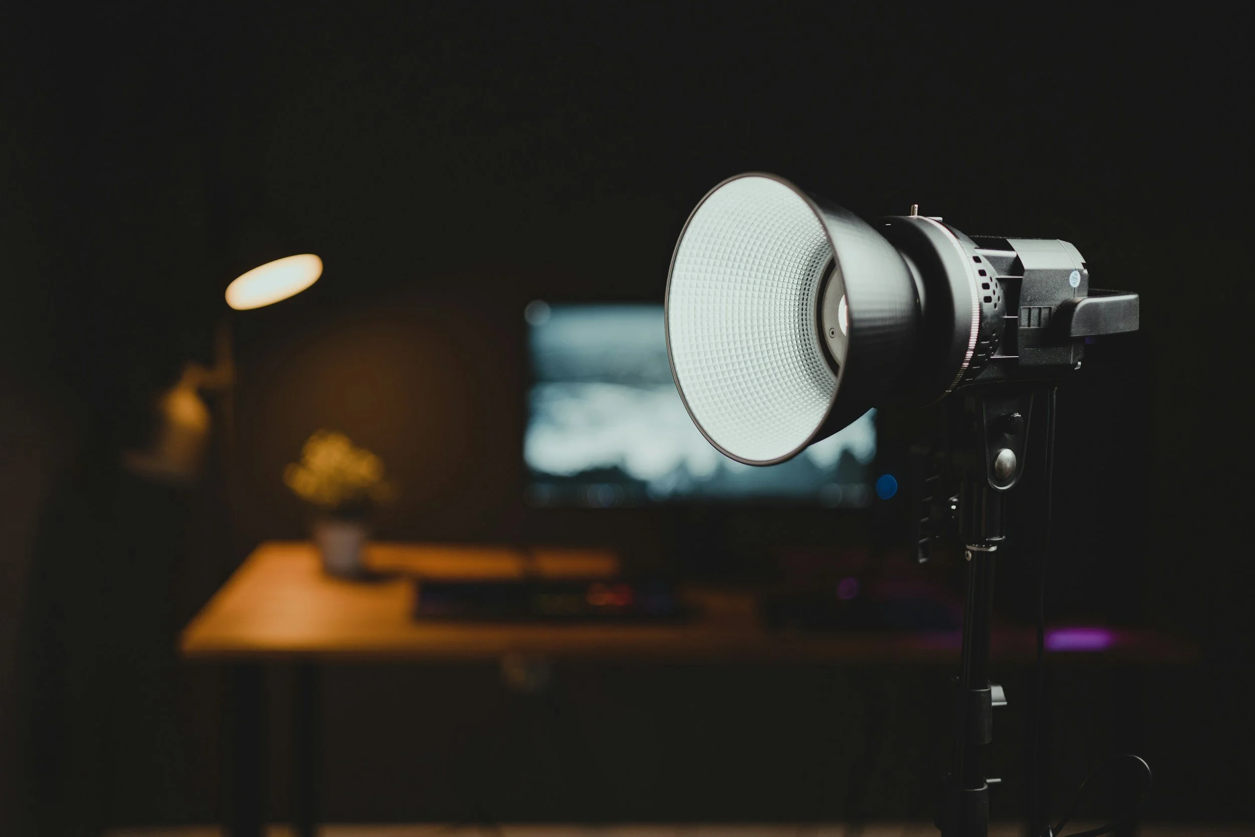 Studio photography setup with a large softbox light and a computer monitor on a desk, with a small potted plant and a desk lamp in the background.