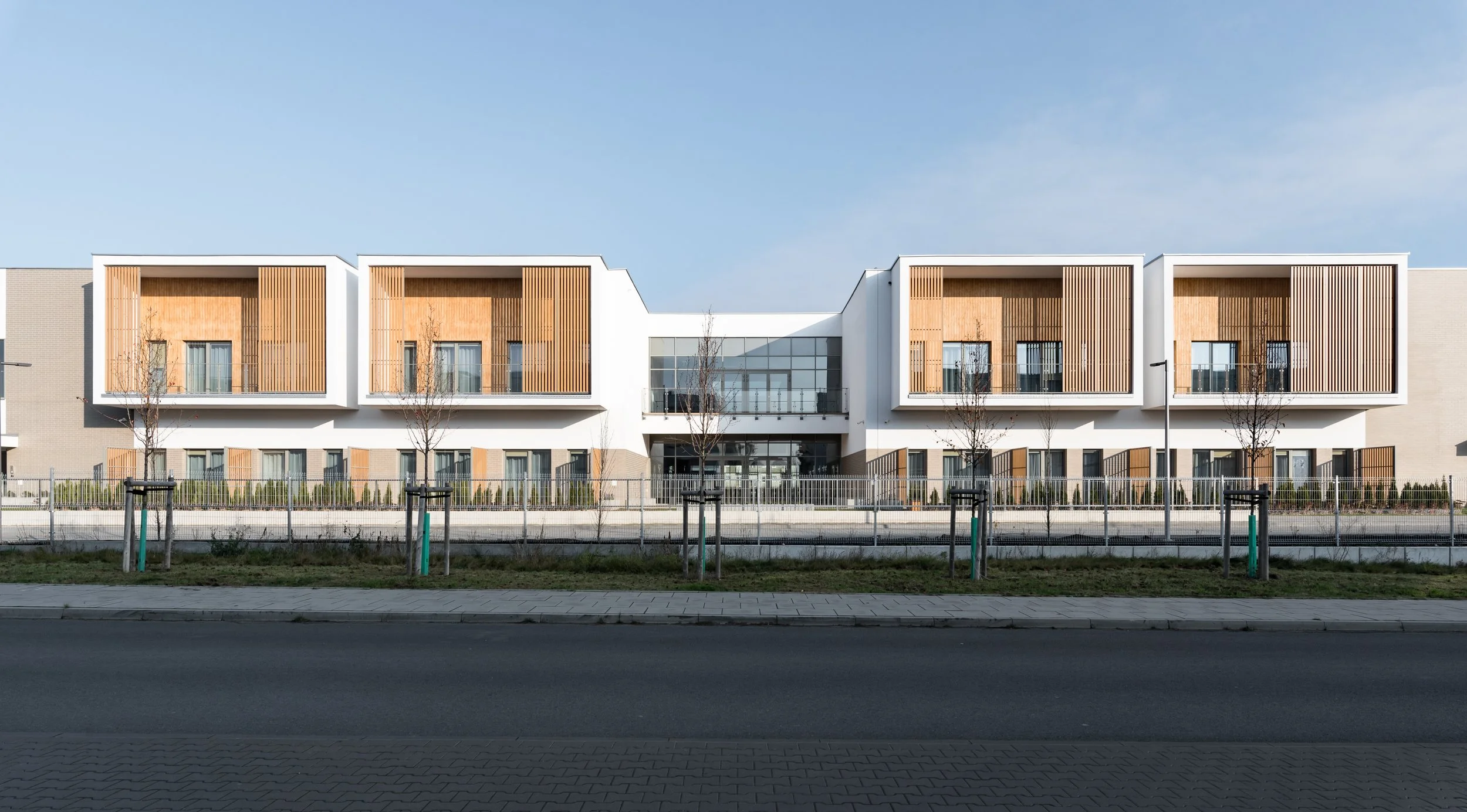 Modern white apartment building with wooden panels, large glass entrance, small trees, and a fence in front.