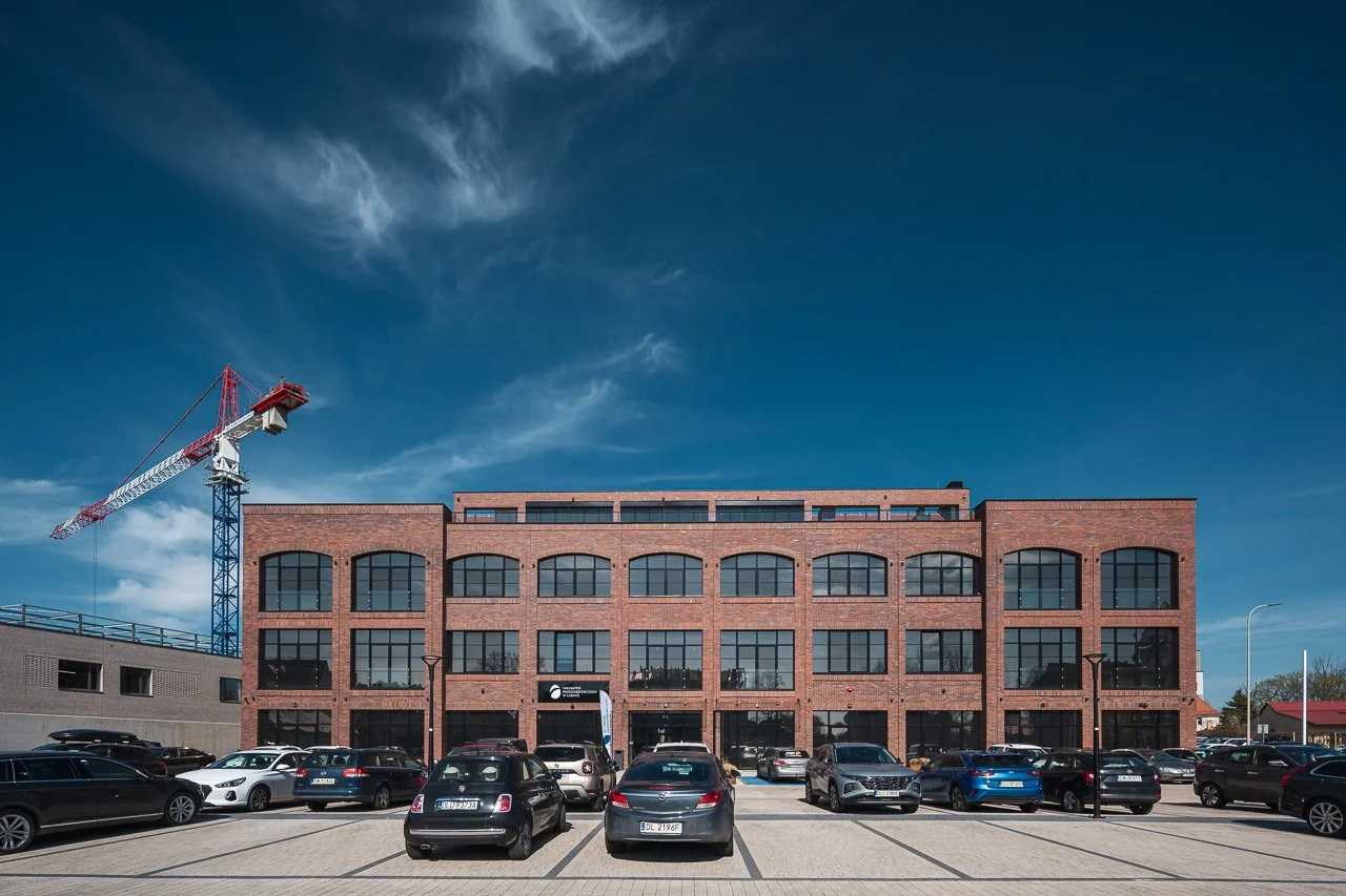 A three-story brick office building with large windows, surrounded by parked cars, under a blue sky with some clouds, and a construction crane on the left.