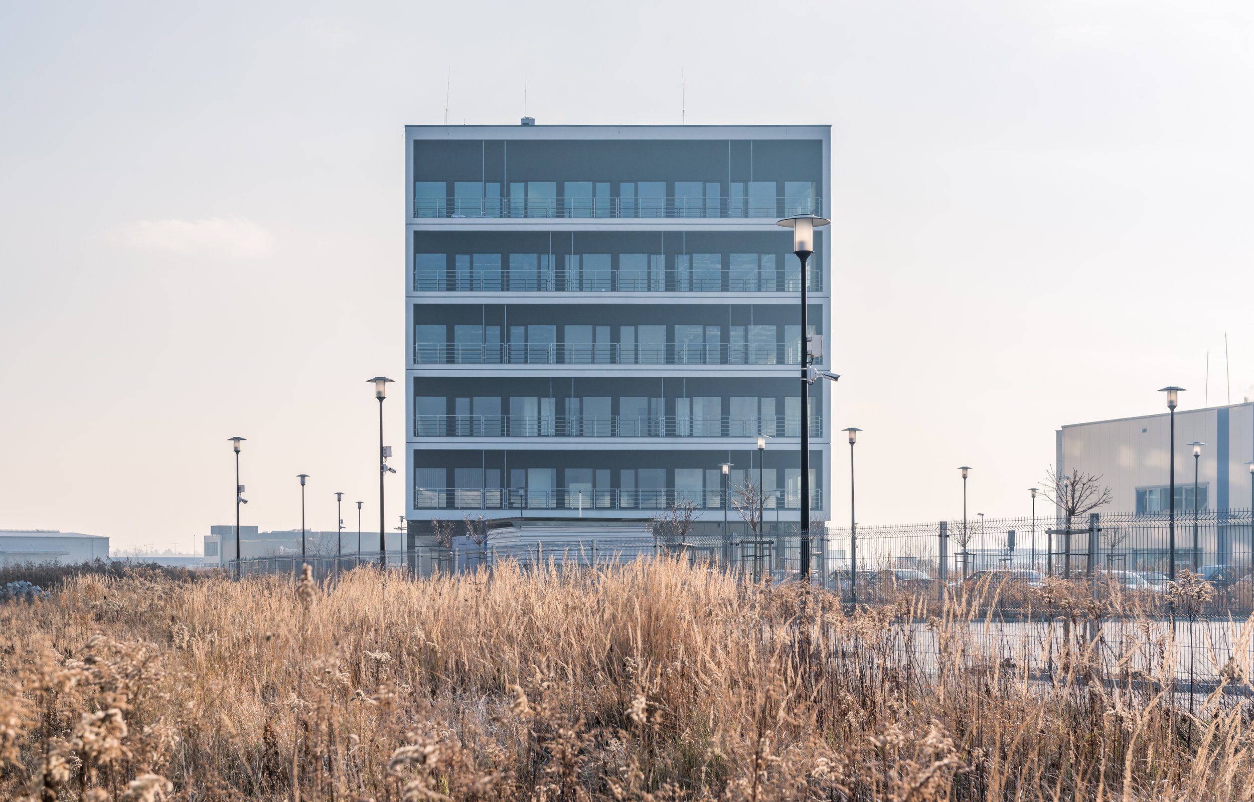 Modern rectangular building with glass balconies and multiple floors, surrounded by a fence, with tall light posts and dry grass in the foreground.