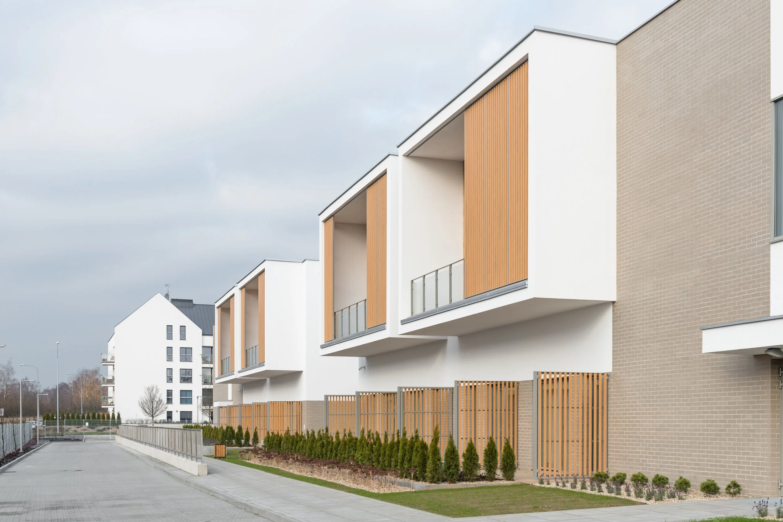 Modern residential building with white walls, wooden balcony accents, and a landscaped sidewalk with small trees and shrubs.