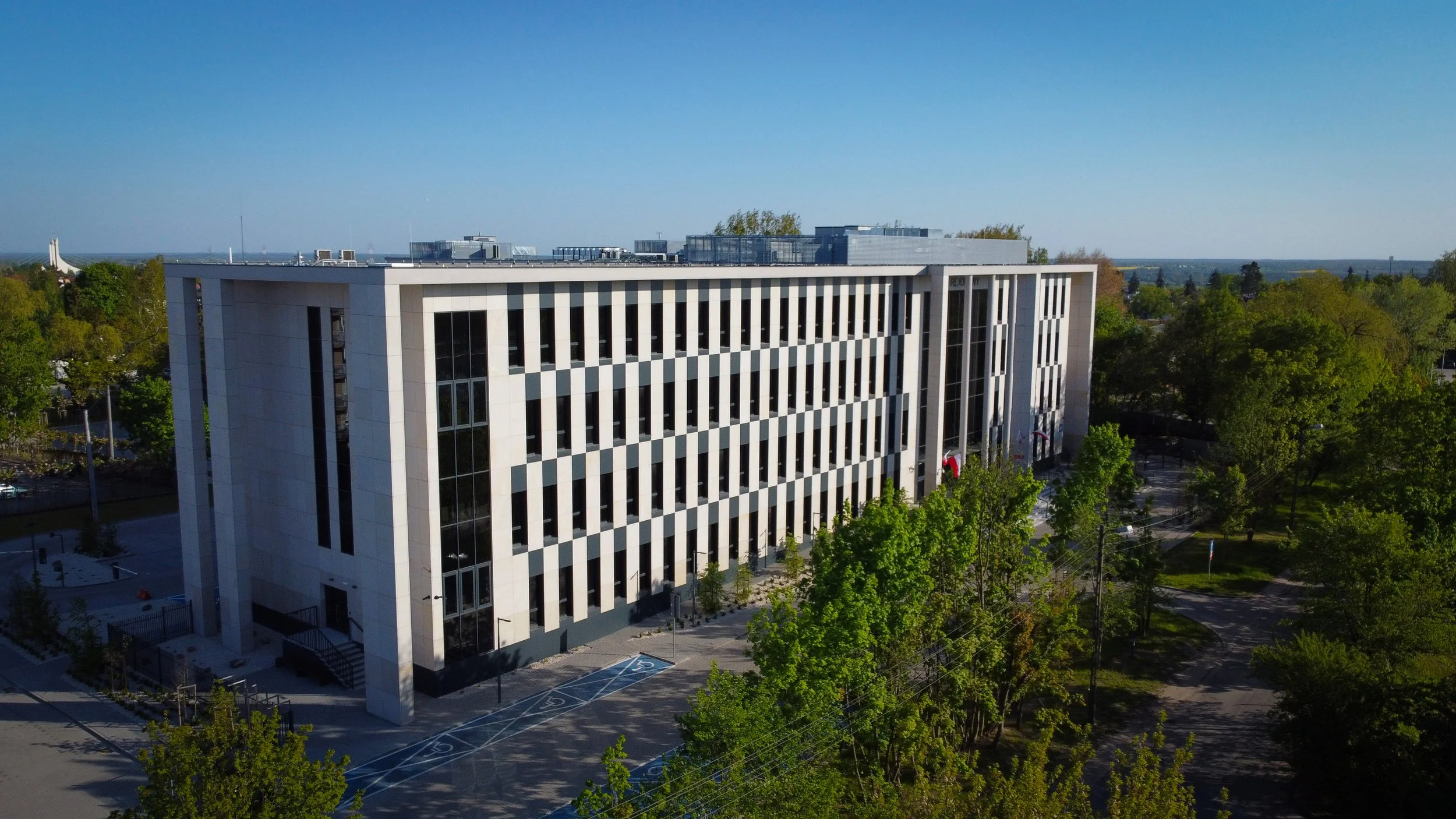 A modern multi-story office building with vertical window strips, surrounded by trees and a parking lot, under a clear blue sky.