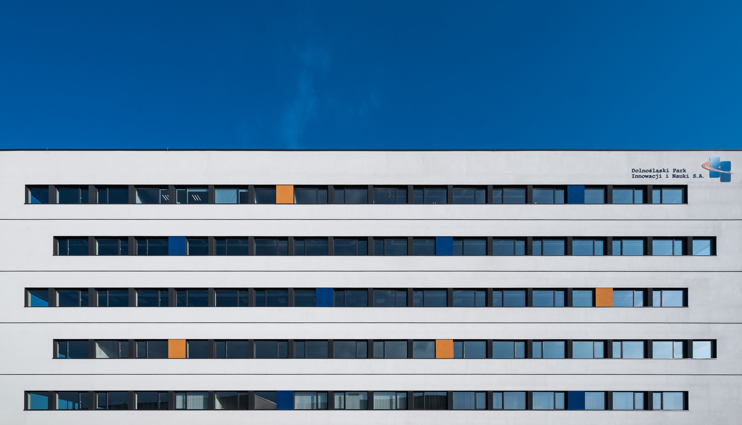 Modern white multi-story building with horizontal rows of windows and colorful orange and blue accents against a clear blue sky.