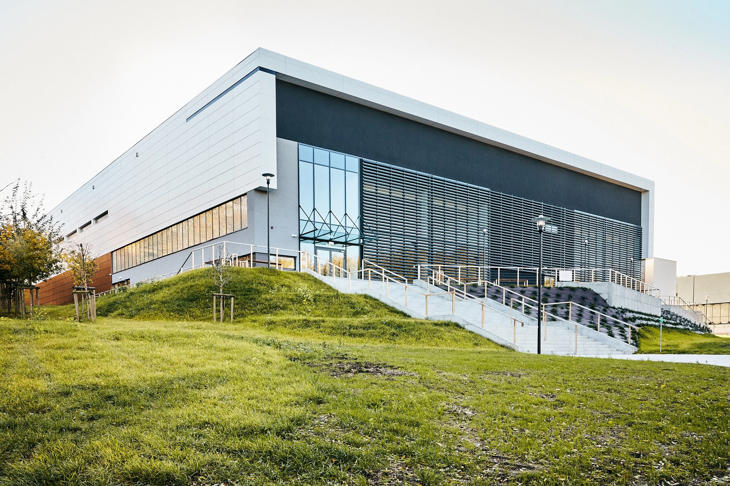 Modern building with large glass windows and slatted exterior, set on a grassy hill with a concrete staircase leading up to the entrance, around sunset.