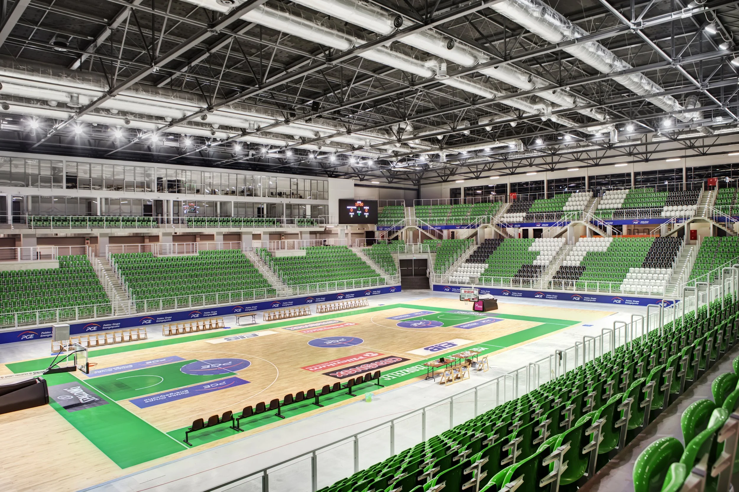 Empty indoor basketball arena with green, white, and black seats surrounding a wooden court, bright lighting, and a scoreboard on the wall.
