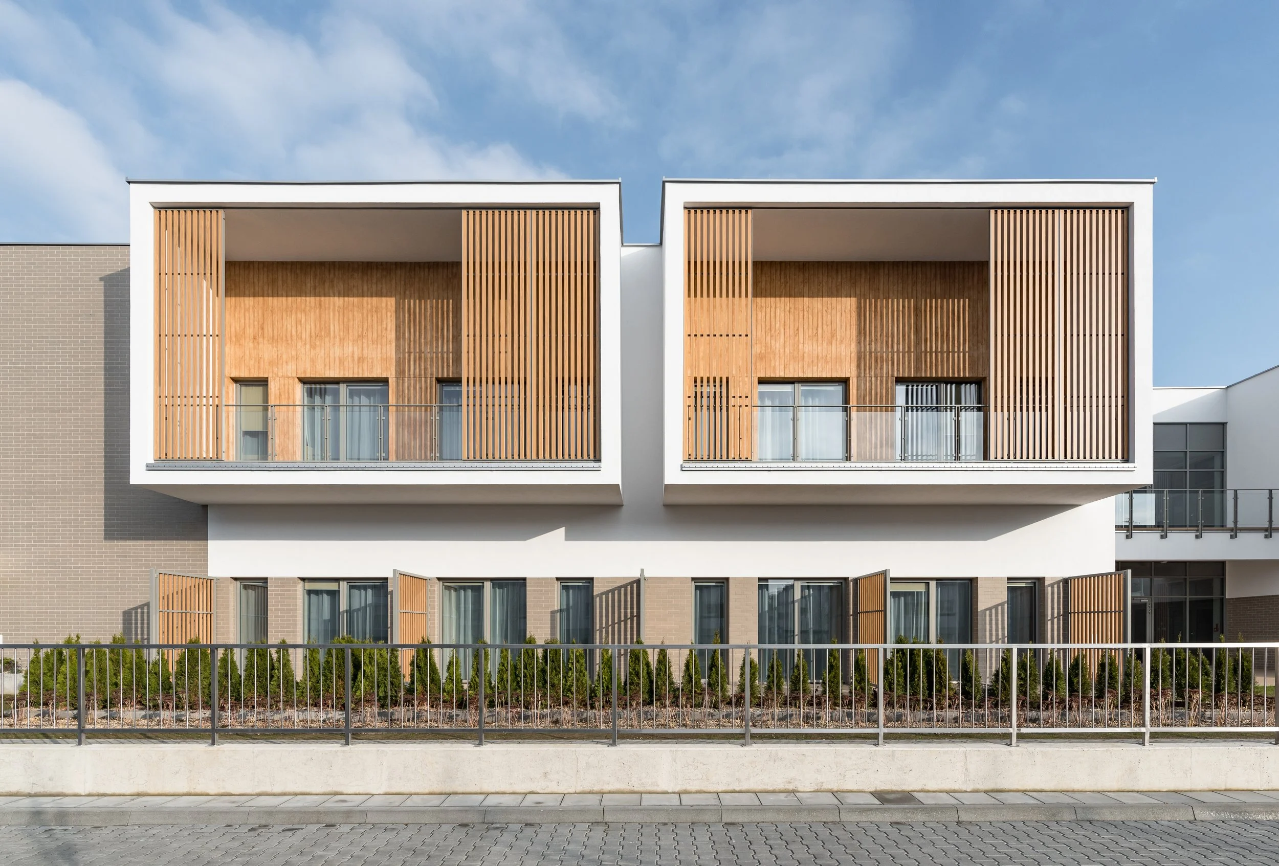 Modern multi-story apartment building with large balconies featuring wooden slat walls, glass railings, and open windows, set against a blue sky with a sidewalk and greenery in the foreground.