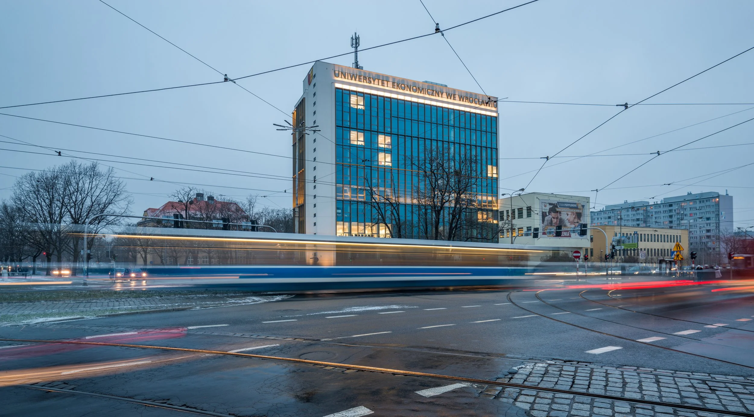 City street scene during twilight with a modern university building labeled "UNIWERSYTET EKONOMICZNY W WROCławiu" in the background. Light trails from passing vehicles and tram tracks visible in the foreground, with leafless trees and additional buil