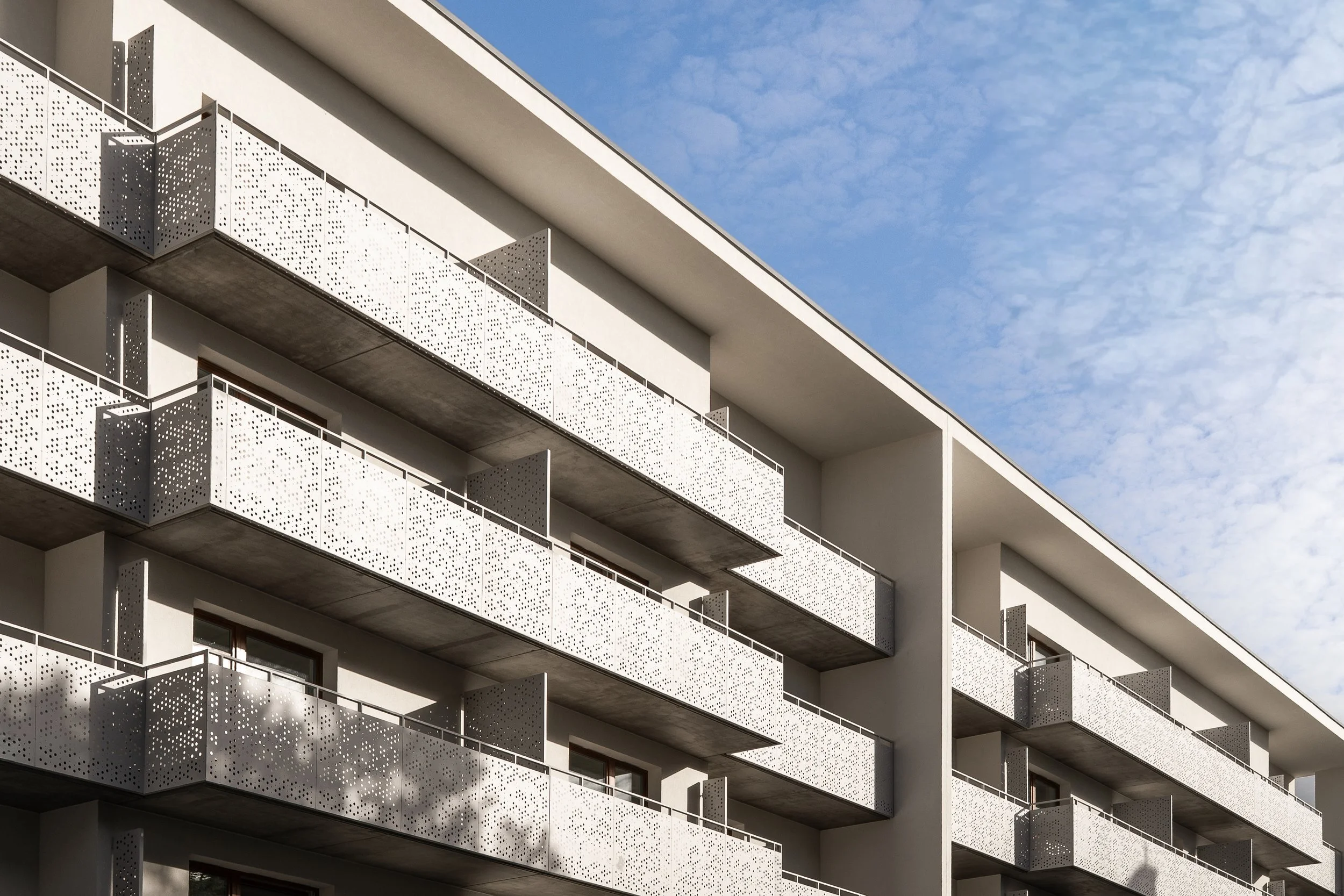 Modern multi-story apartment building with white perforated metal balcony railings, dark balcony floors, and a blue sky with wispy clouds in the background.