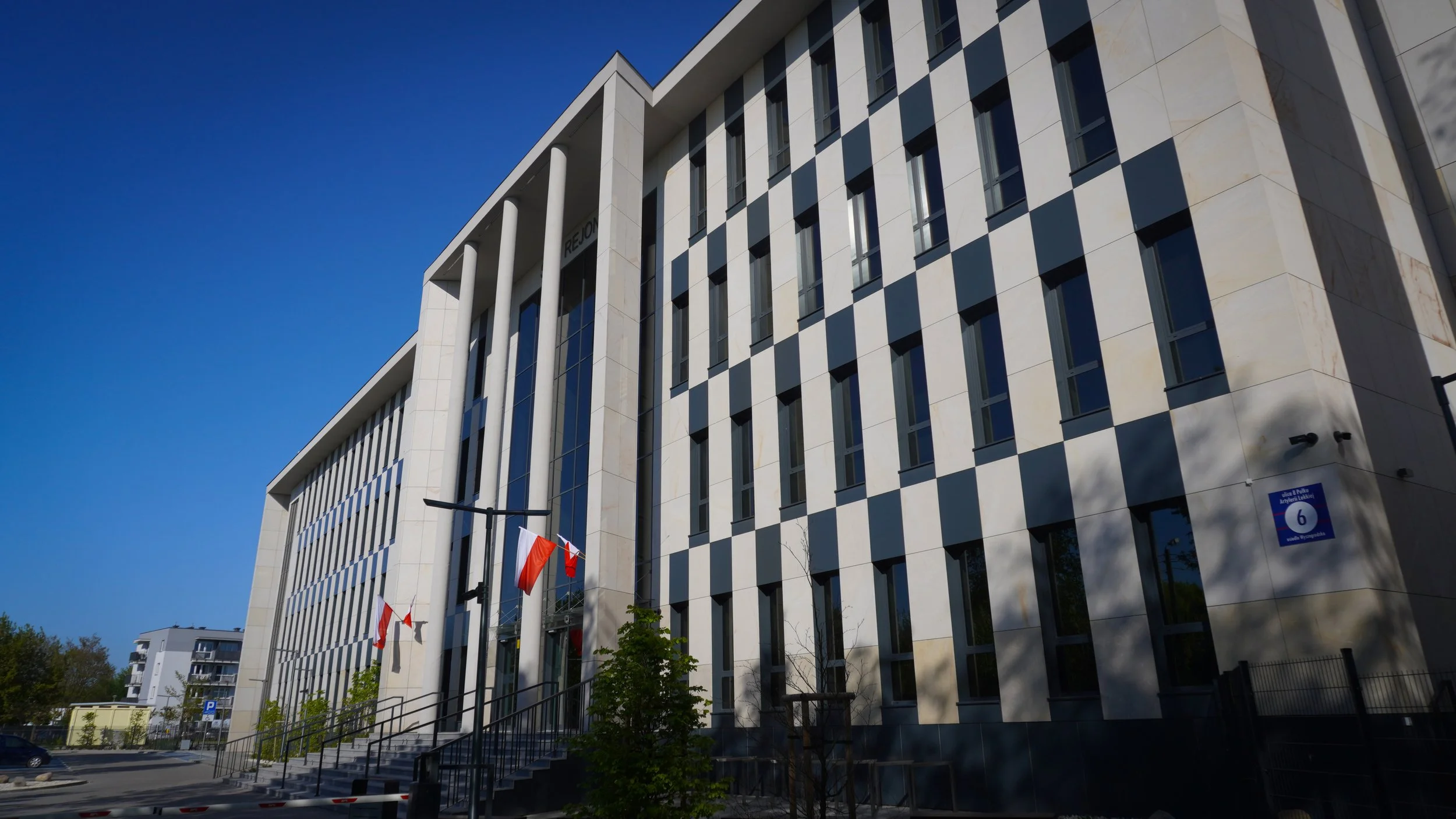 Modern white building with vertical windows and checkered black and white wall panels, flags flying and stairs leading up to the entrance, under a clear blue sky.