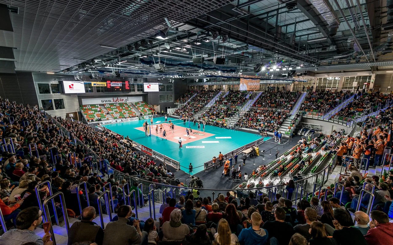Indoor volleyball match with players on the court, surrounded by an audience in the stands, in a large sports arena.