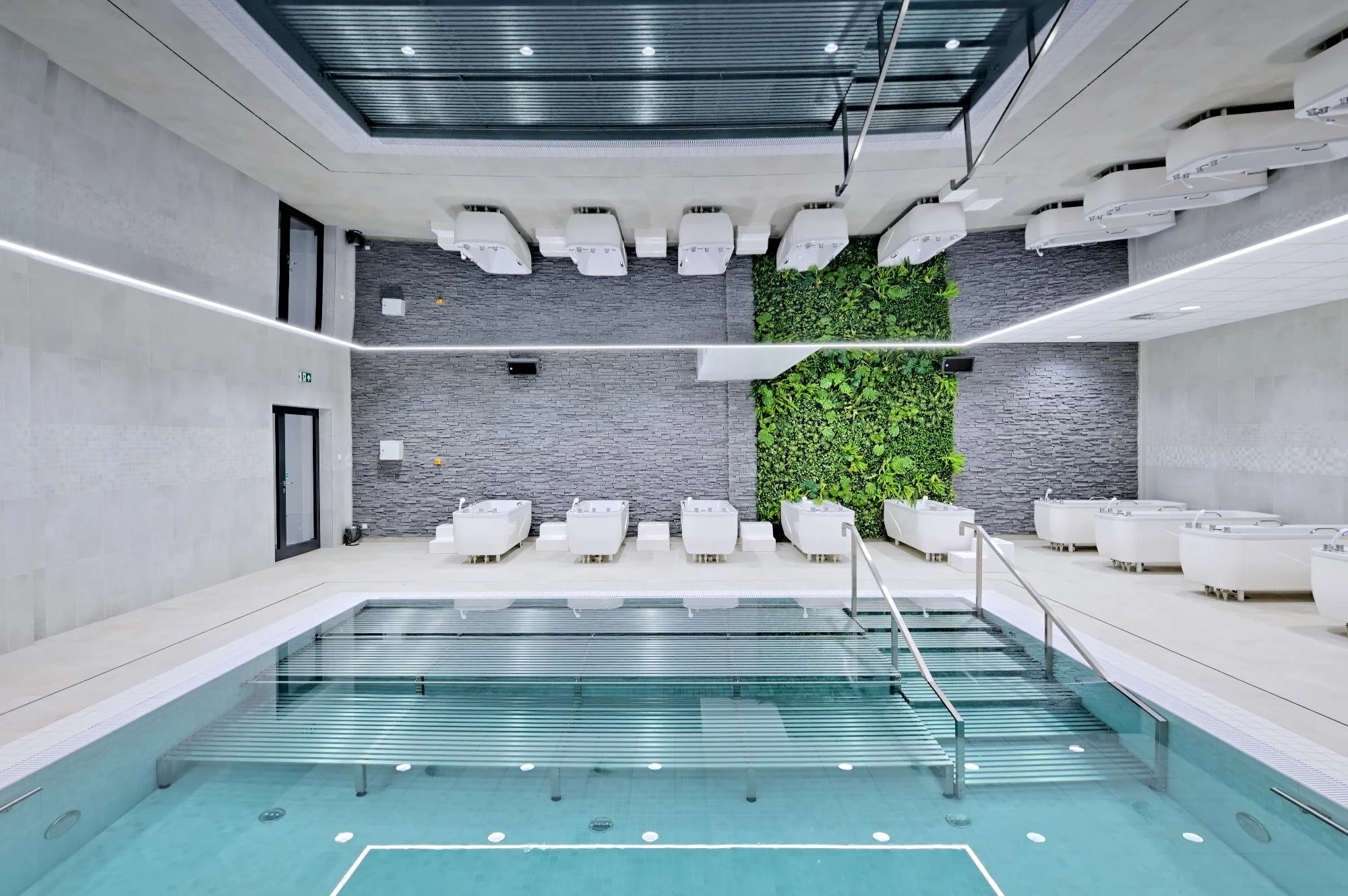 Indoor pool area with a glass pool gate, white whirlpool tubs, and a green plant wall.