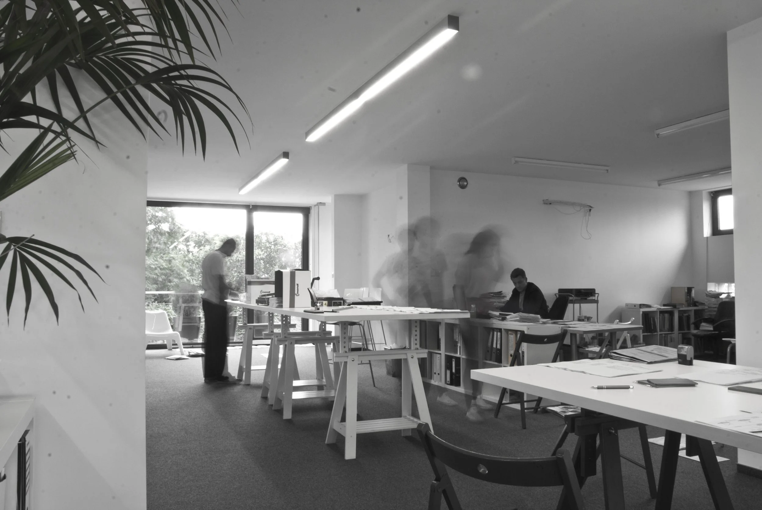 An office with several desks and chairs. There are three people in the office, with two appearing as blurred motion figures, and one person sitting at a desk. Large windows let in natural light, with blinds partially drawn. Office supplies and documents are scattered across the desks.