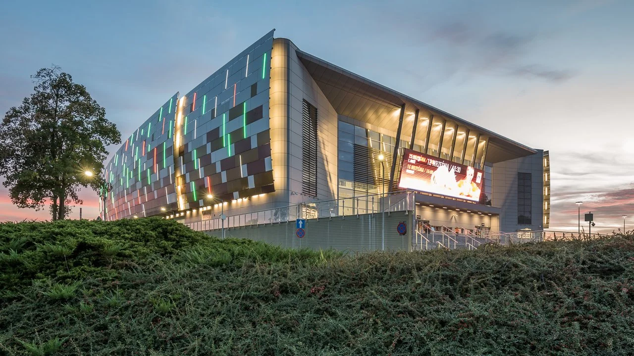Modern stadium with illuminated facade and digital billboard at sunset surrounded by greenery.
