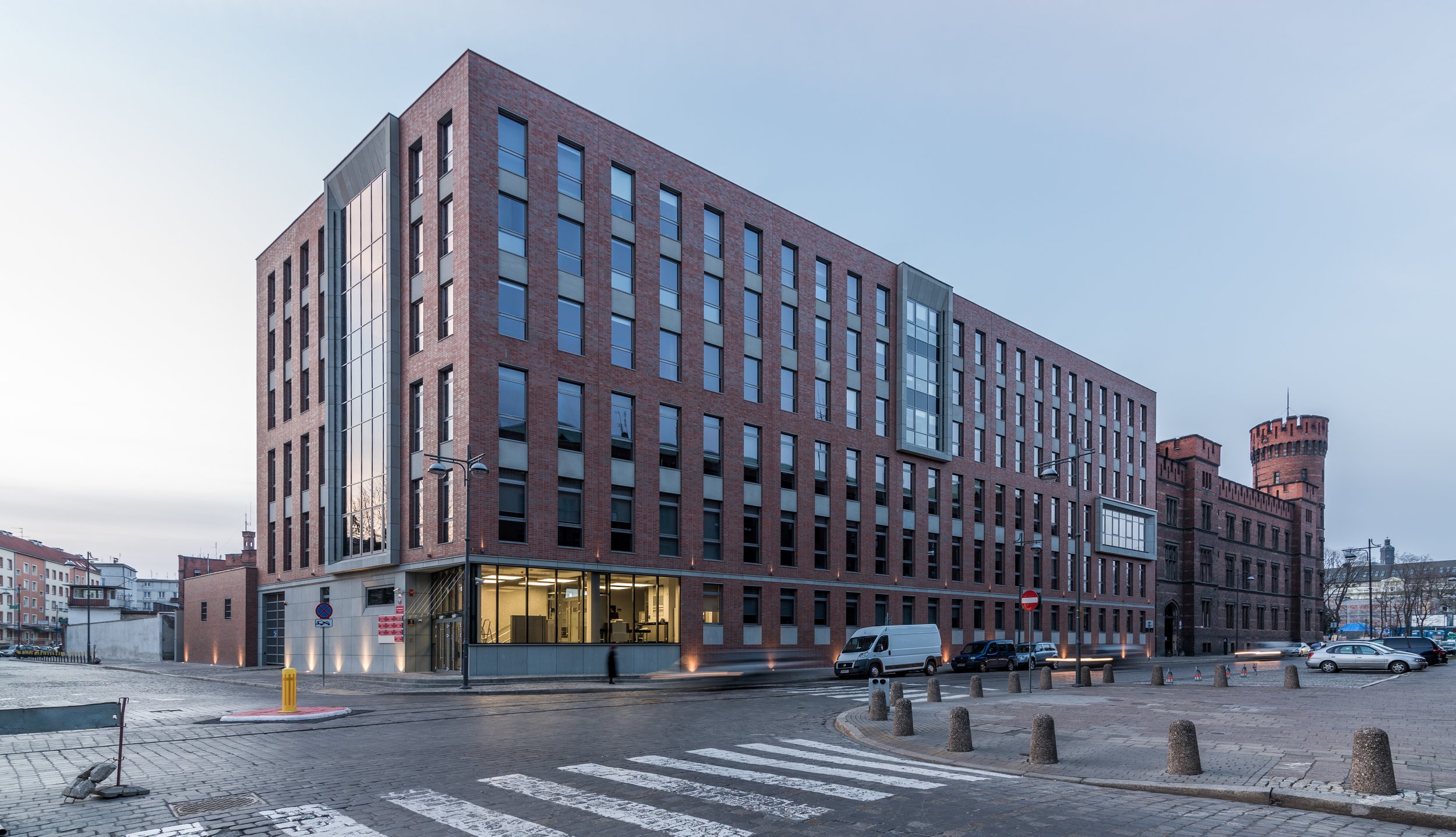 Modern brick office building with large glass windows, located on a city street with cars parked in front, and a historic tower in the background.