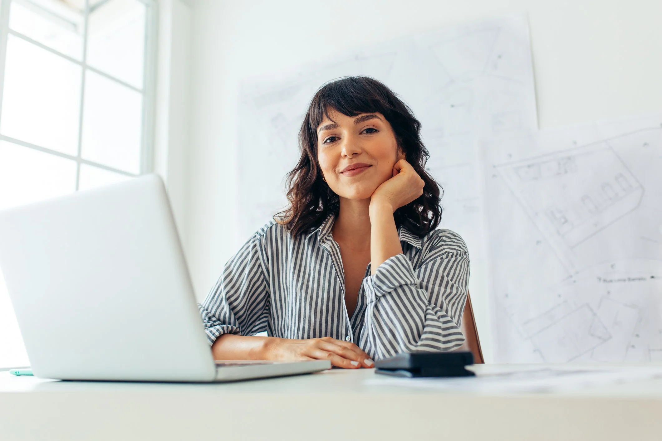 Women sitting at a desk in an office, smiling, with a laptop and a smartphone on the desk, and architectural plans on the whiteboard behind her.