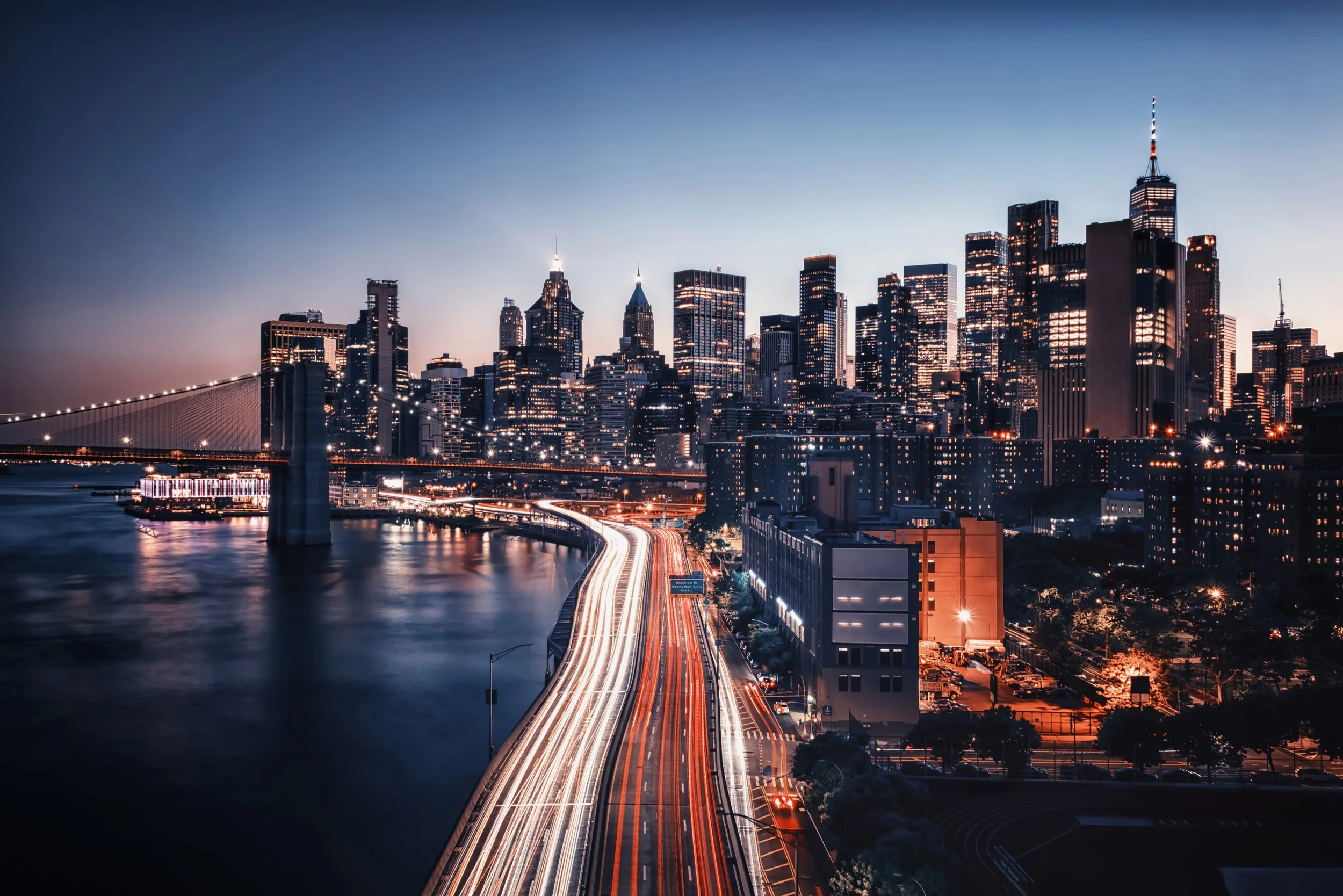 Night view of a city skyline with tall buildings, bridges, and a river, illuminated with city lights.